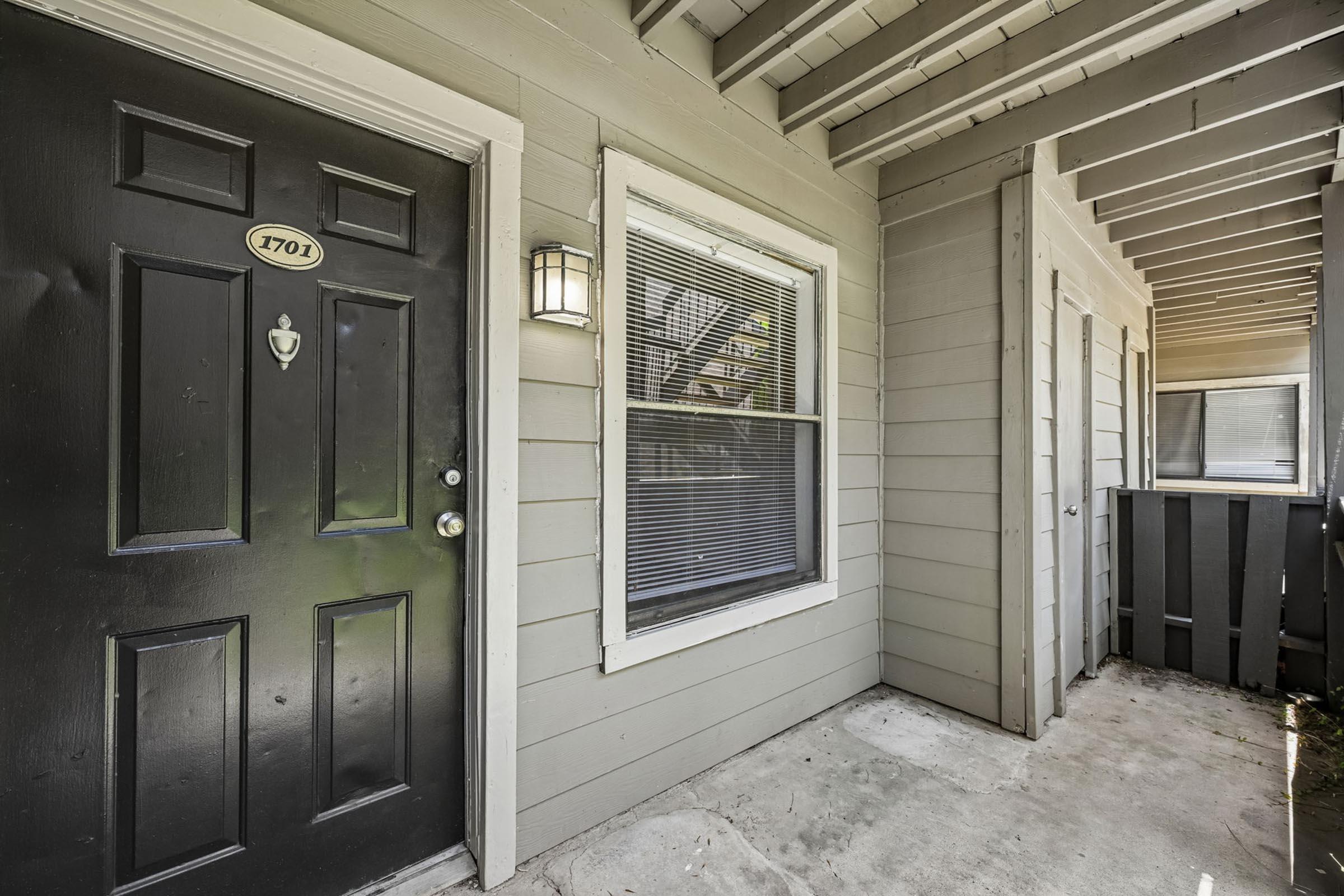 Exterior view of an apartment entrance featuring a dark door with a number plaque labeled "101," a wall-mounted light fixture, and a window beside the door. The surrounding walls are painted in light gray, with a covered porch area and a visible railing leading to the adjacent unit. The ground is concrete and slightly weathered.