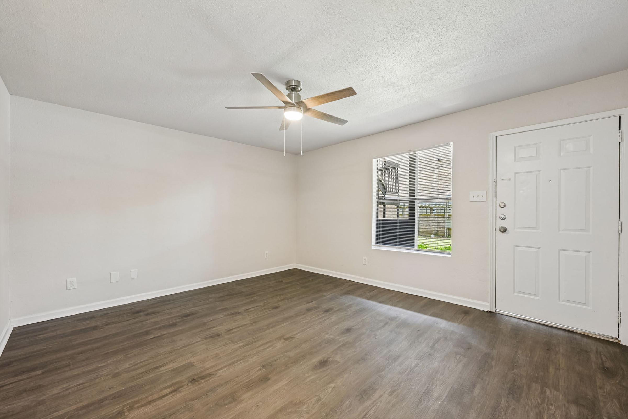An unfurnished room featuring light brown laminate flooring, a ceiling fan with light fixture, and a window allowing natural light. A front door is visible on the right side, and plain white walls create an open, airy atmosphere. The space is suitable for various design options.
