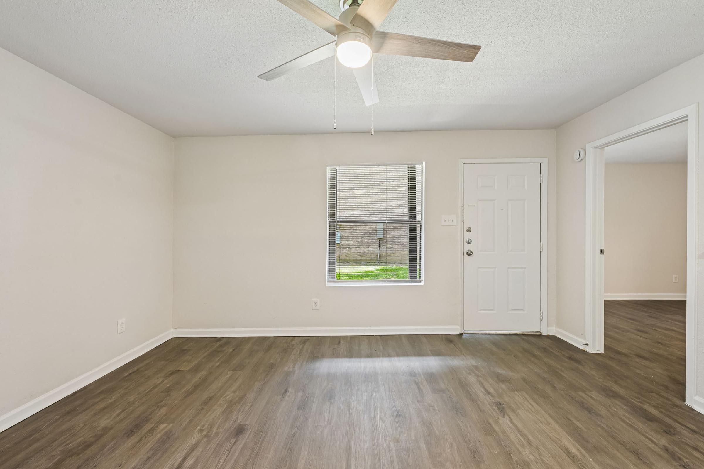 A well-lit living room featuring a ceiling fan, light-colored walls, and new laminate flooring. A window allows natural light to enter, and the entrance door and hallway are visible. The space appears clean and inviting, ready for furniture placement.