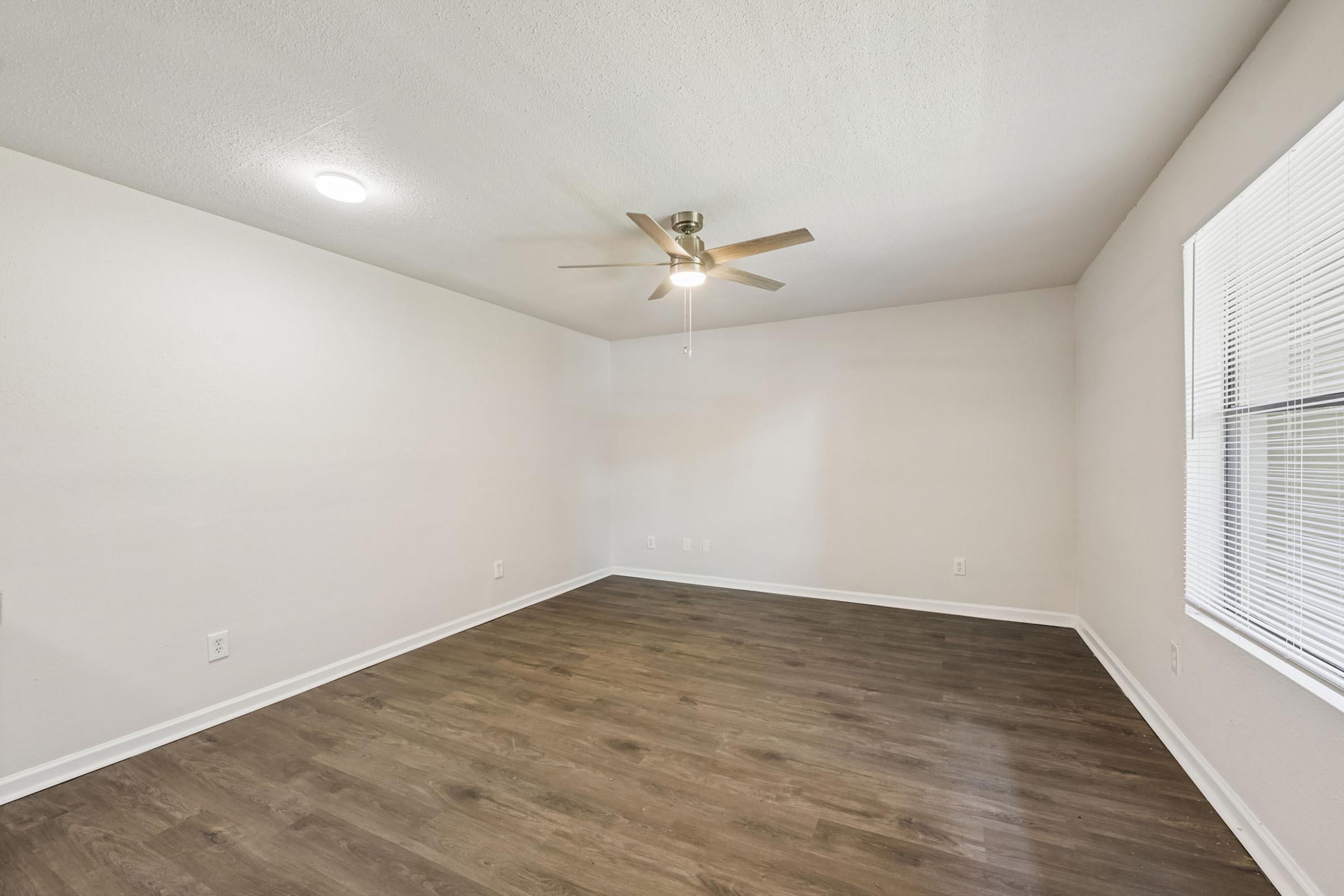 A vacant room featuring light-colored walls, a ceiling fan, and large windows with blinds. The floor is made of wood-like flooring, and the space is bright and airy, offering a clean and open layout suitable for various furniture arrangements.