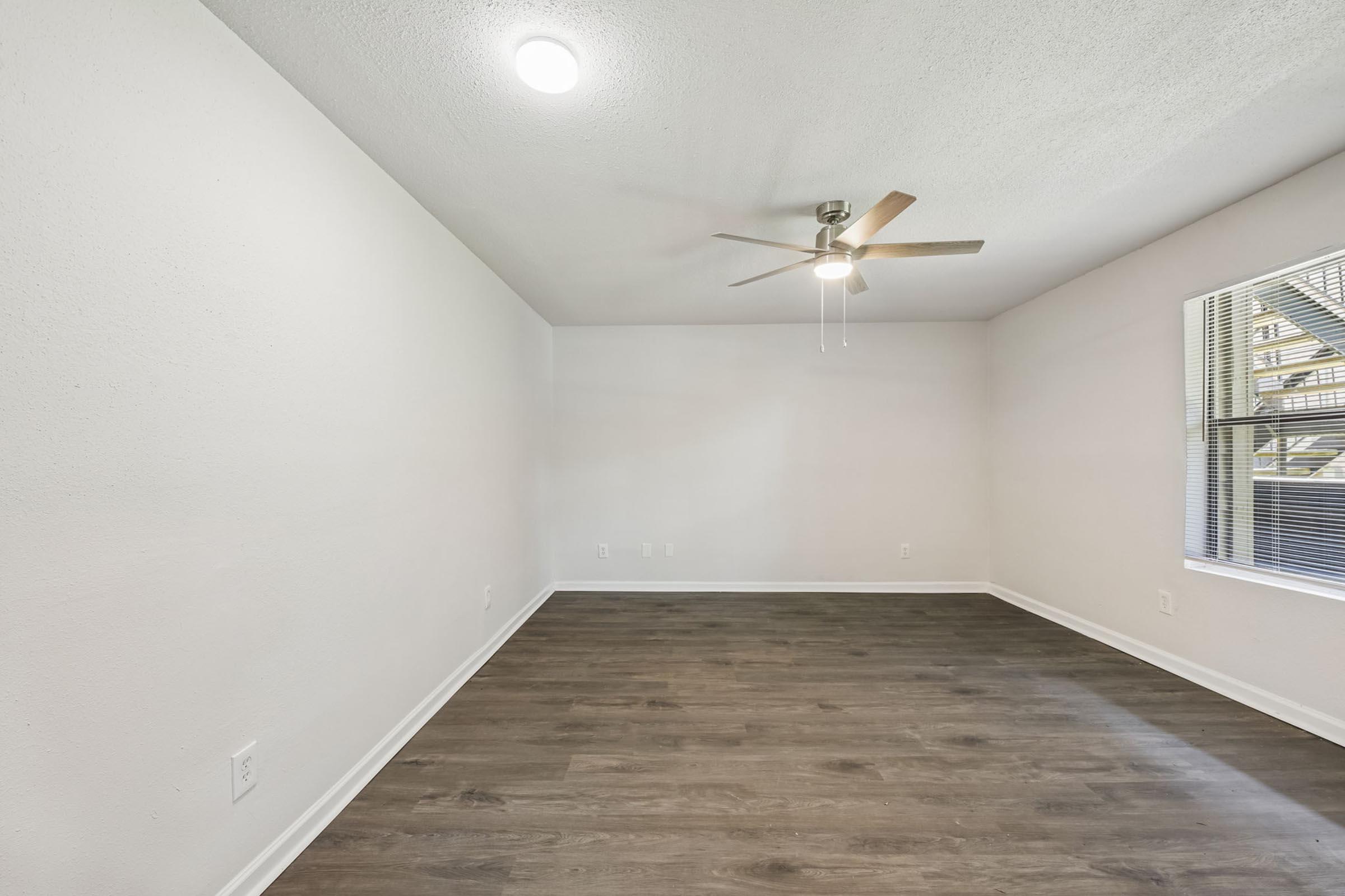 A clean, empty room with light-colored walls and a ceiling fan. The floor is a brown laminate, and there is a window on the right side allowing natural light to enter. The space appears bright and ready for furnishing.