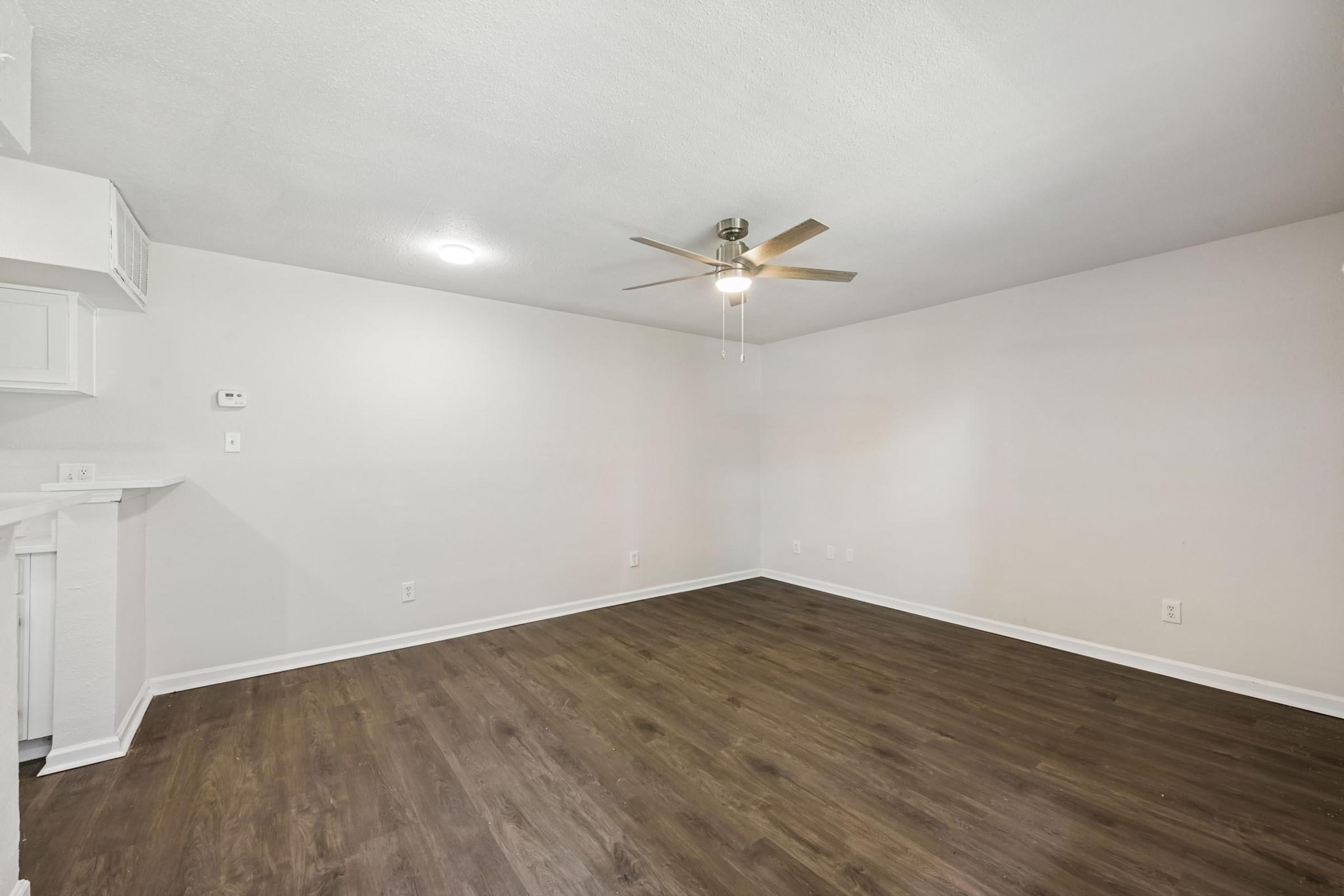 A spacious, empty room with light-colored walls and a ceiling fan. The floor is dark wood, and there are no furnishings. A small area on the left suggests a kitchenette with white cabinetry. Soft, diffused lighting from ceiling fixtures illuminates the space.