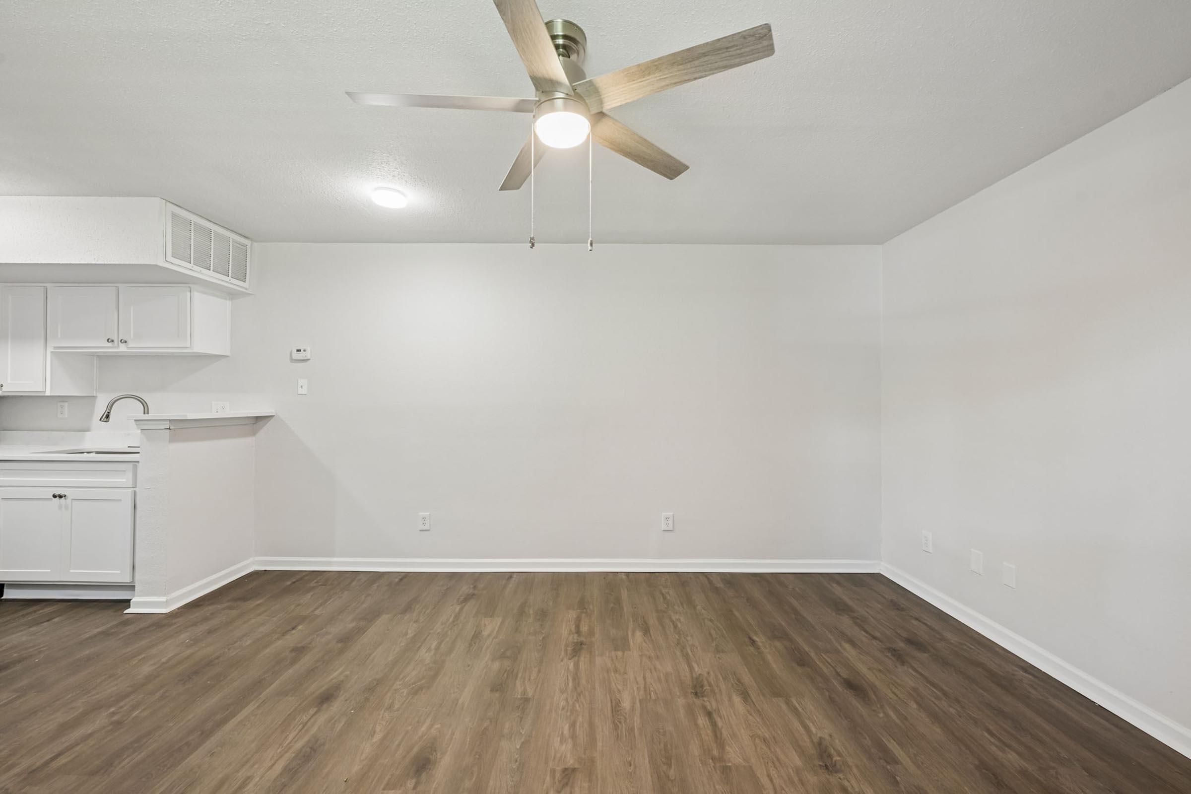 A clean, spacious interior of an apartment or room with a ceiling fan and light fixture. The walls are painted white, and there is a wood-like laminate floor. In the background, there is a small kitchen area with white cabinetry, but the space appears empty and ready for furniture.