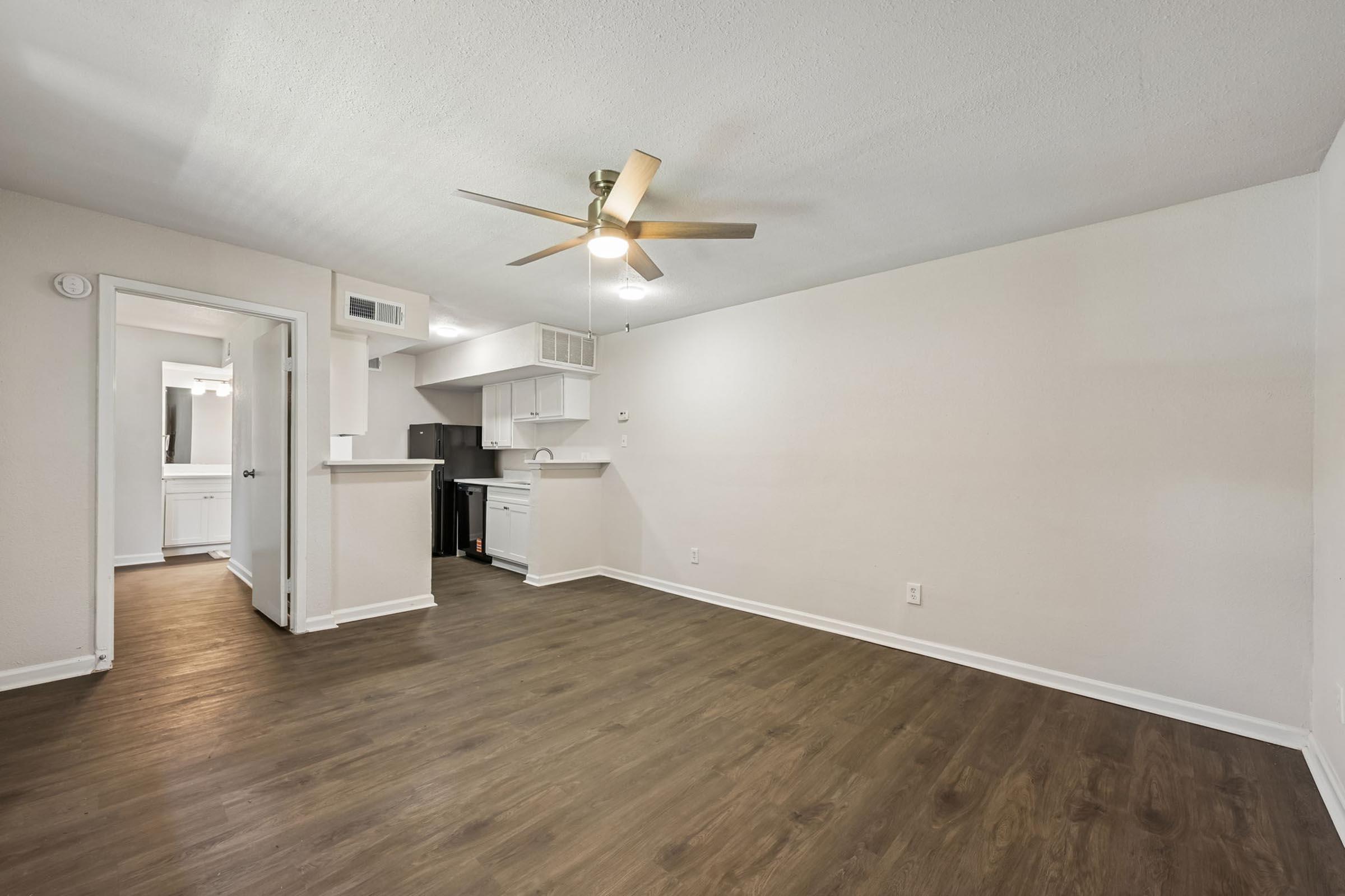 Spacious living area with light-colored walls and a ceiling fan. The floor is made of wood-like laminate. A doorway leads to the kitchen, featuring cabinets and appliances. Natural light comes through a window, enhancing the open atmosphere.
