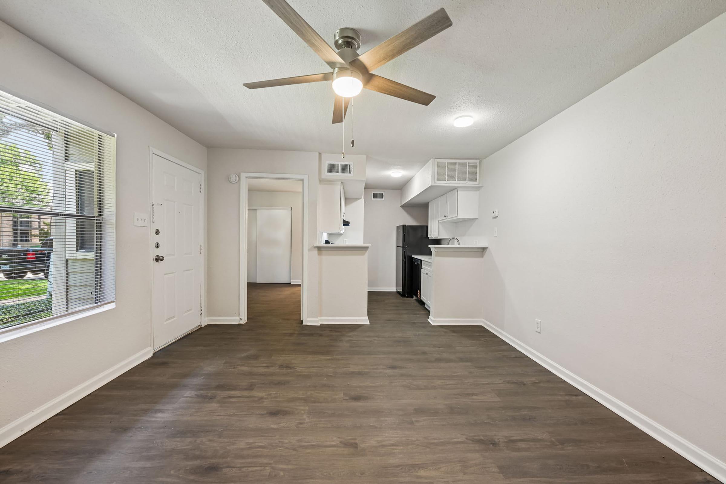 Interior view of a modern apartment living area showcasing a ceiling fan, light-colored walls, and a spacious layout. The room features large windows allowing natural light, hardwood-style flooring, and an open kitchen area with white cabinetry.