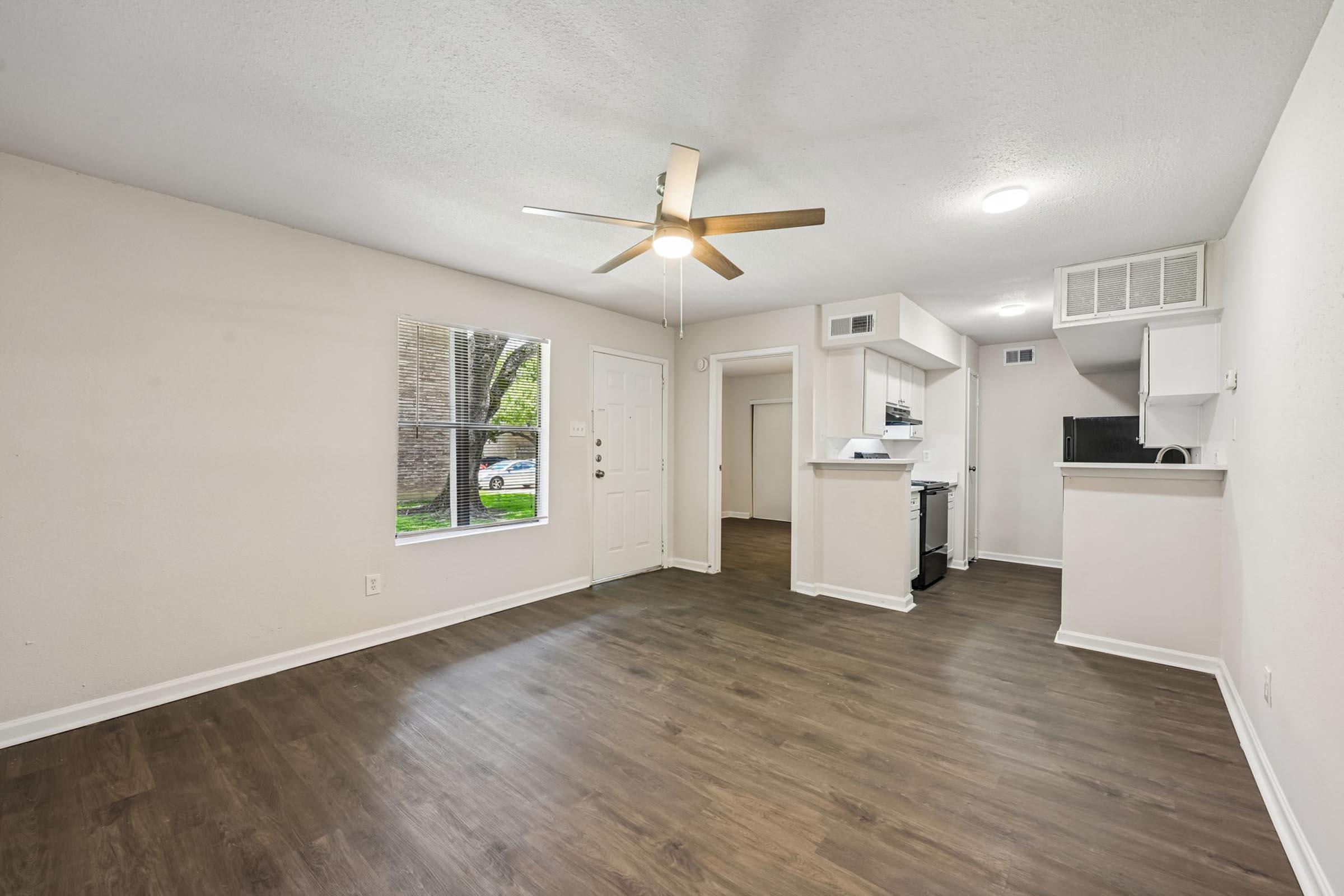 Cozy living space featuring a light-colored wall, a ceiling fan, large window allowing natural light, and modern vinyl flooring. The room connects to a kitchen with a bar area, equipped with white cabinetry and appliances, creating an open-concept layout perfect for relaxation and entertaining.