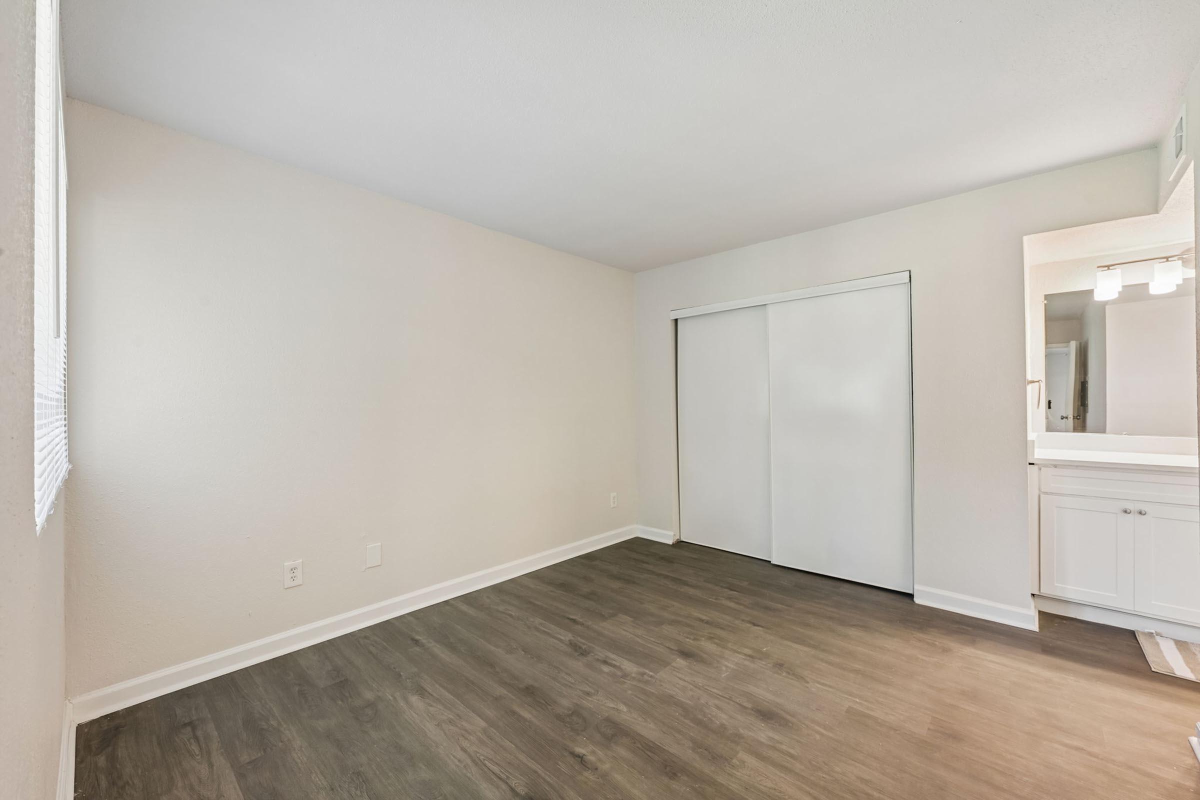 A vacant room with light-colored walls and a smooth, dark wood-style floor. There is a large sliding closet door on one side, and a doorway leading to a bathroom area on the opposite side, featuring a large mirror and light fixtures. Natural light enters through a window.