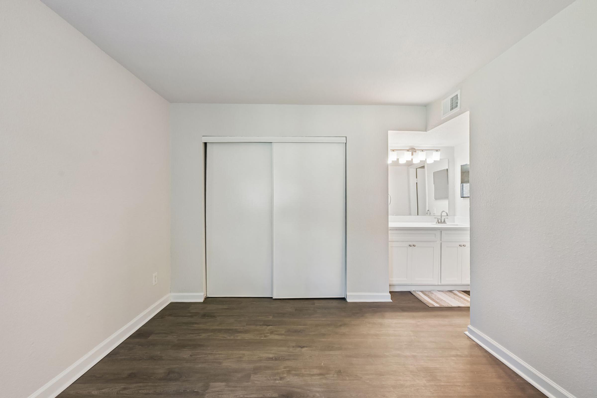 A bright, empty room featuring light-colored walls and a large closet with sliding white doors. The floor is made of dark wood, and there is a doorway leading to a bathroom area with modern fixtures and lights visible in the mirror. The overall ambiance is clean and spacious.