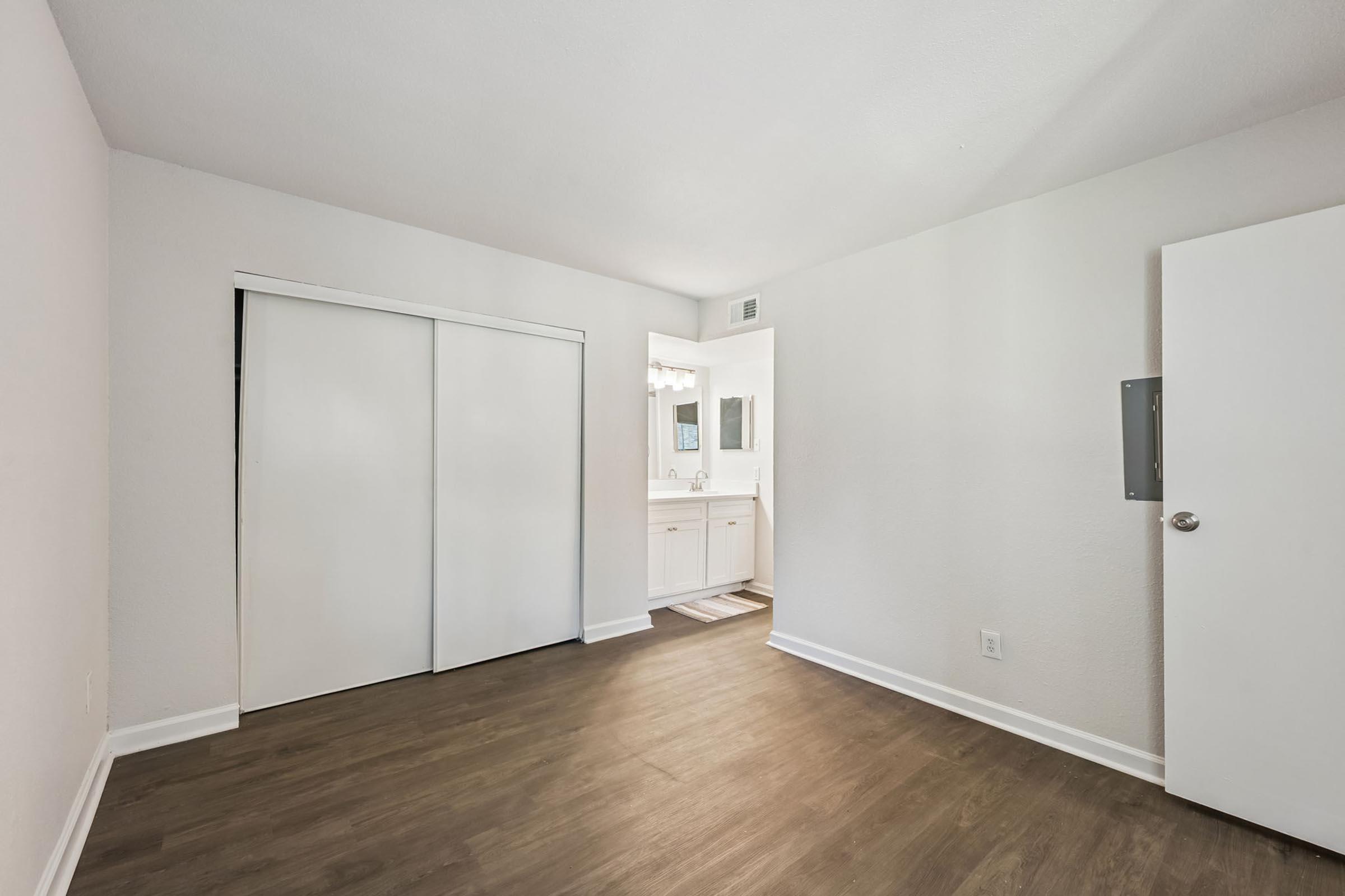 A spacious, empty room with light-colored walls and a wooden floor. There are two closet doors on one side and a doorway leading to a bathroom area with a sink and mirror visible in the background. Natural light illuminates the space, creating a welcoming atmosphere.