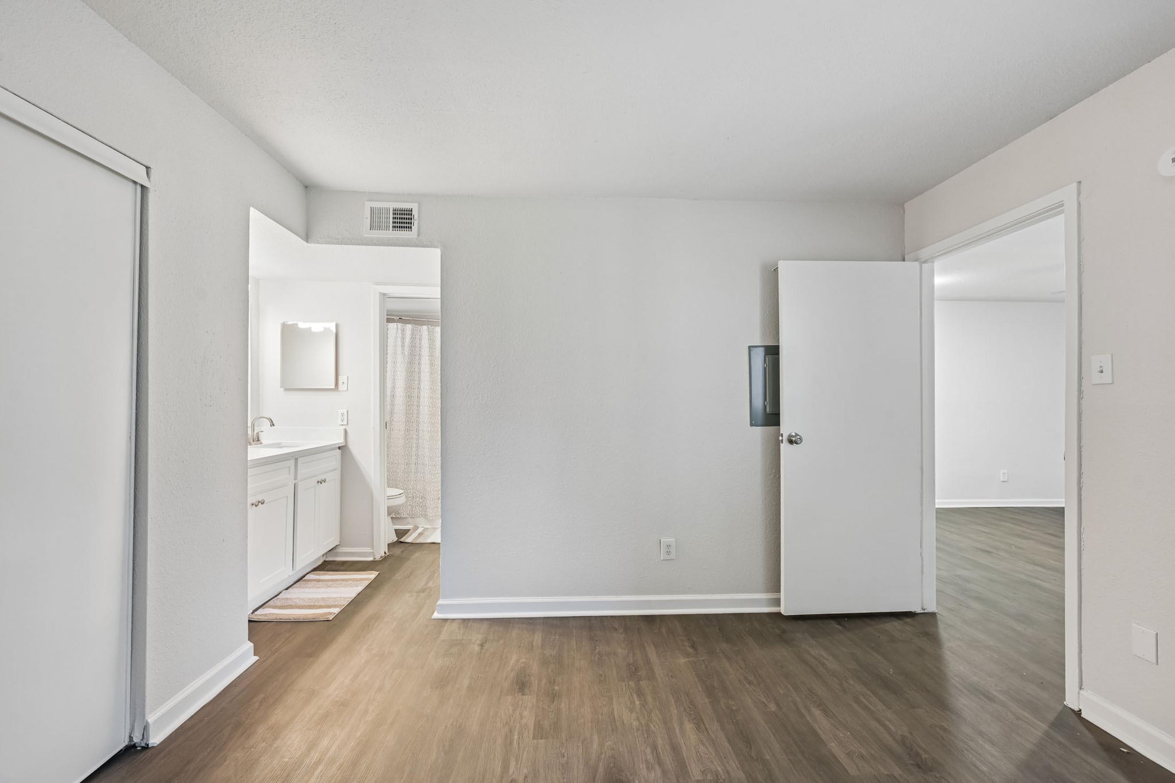A well-lit interior space featuring a light gray wall, a brown laminate floor, and open doorways leading to a bathroom and another room. The bathroom entrance has a mirror and a brightly lit vanity area visible. The room appears clean and inviting with natural light.