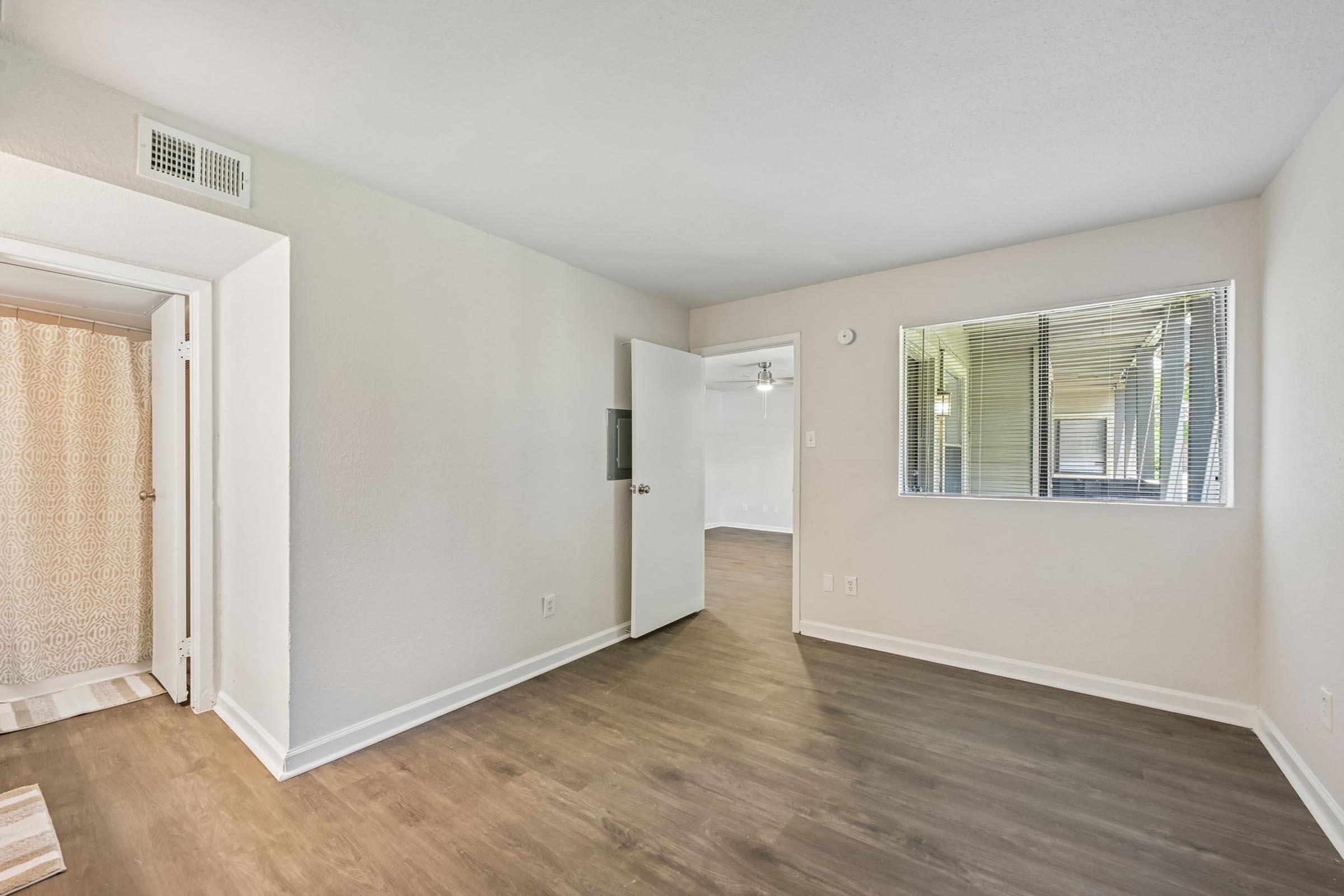 A light-filled, modern living space featuring hardwood-style flooring, neutral walls, and a large window. On one side, there is a small bathroom area with a shower curtain. The room is designed with a minimalist aesthetic, making it suitable for various interior styles.