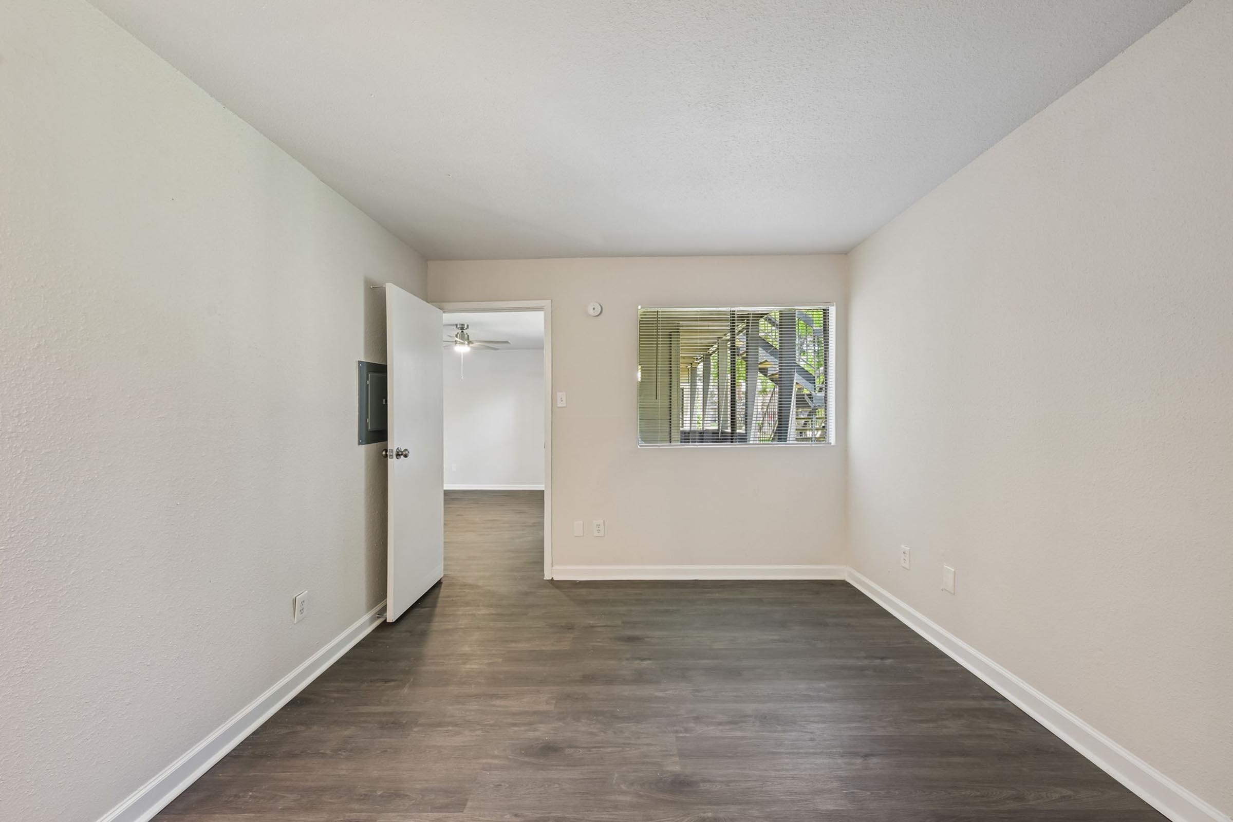 A small, empty room with light-colored walls and a dark wooden floor. There is an open door on the left leading to another space, and a window on the right allowing natural light. The overall atmosphere is bright and minimalistic, suitable for various uses.
