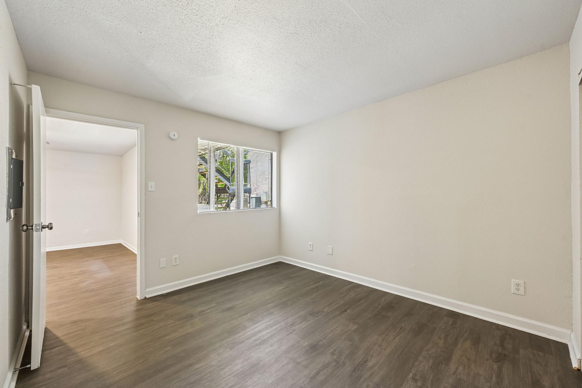 An empty room featuring light-colored walls and a smooth, dark wooden floor. A window on one wall lets in natural light, and there's an open door on the left leading to another room. The space is clean and minimalistic, ideal for furnishing and personalization.