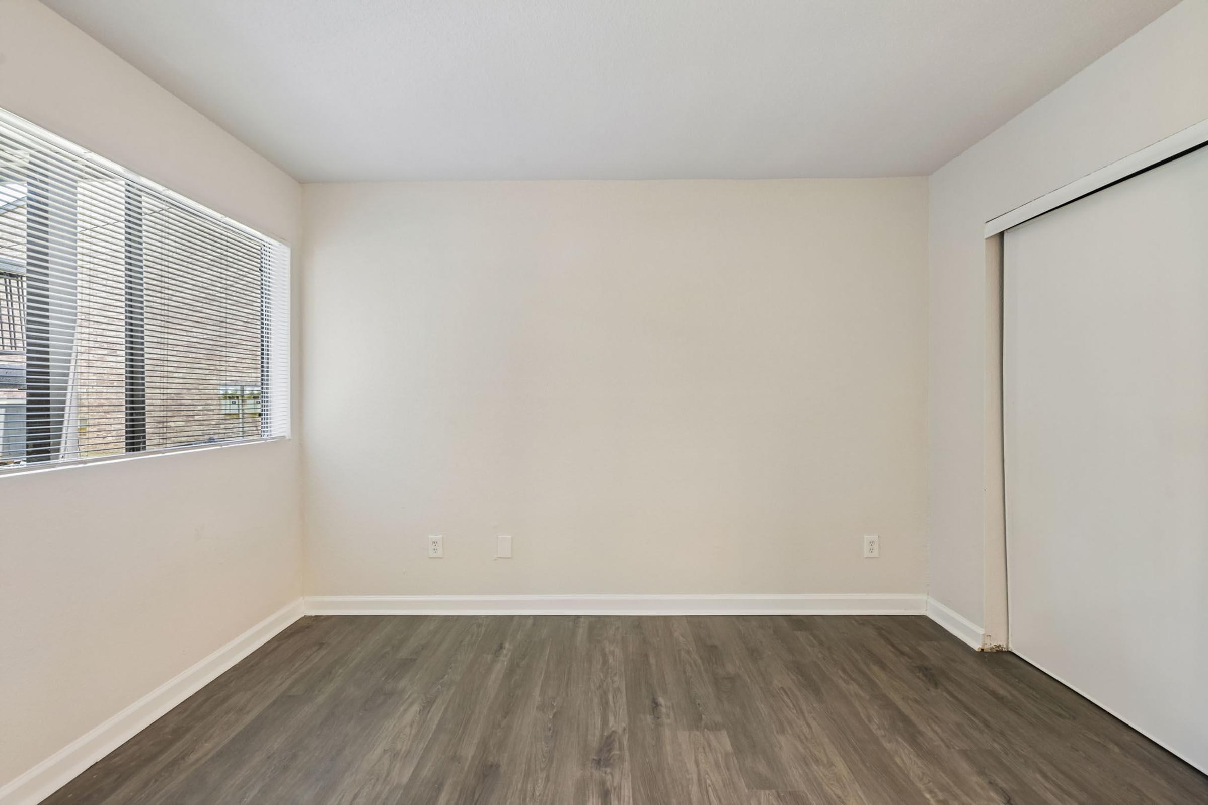 A well-lit, empty room with light-colored walls and a window on the left. The flooring is a modern wood style, and there is a closet door on the right. The space appears clean and ready for furnishing.