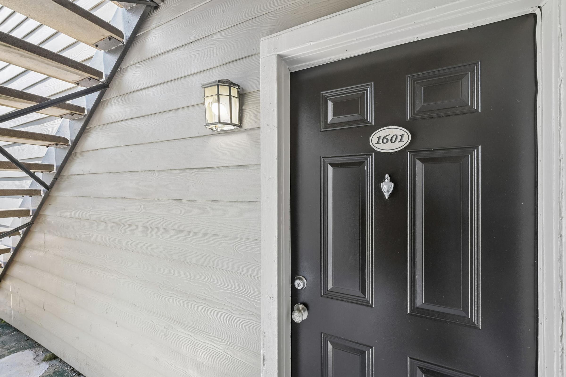 A black front door with the number "1601" displayed on it, featuring a doorbell and a door knocker. A light fixture is mounted beside the door on a light-colored wooden wall. A staircase is visible in the background, leading up beside the entrance.