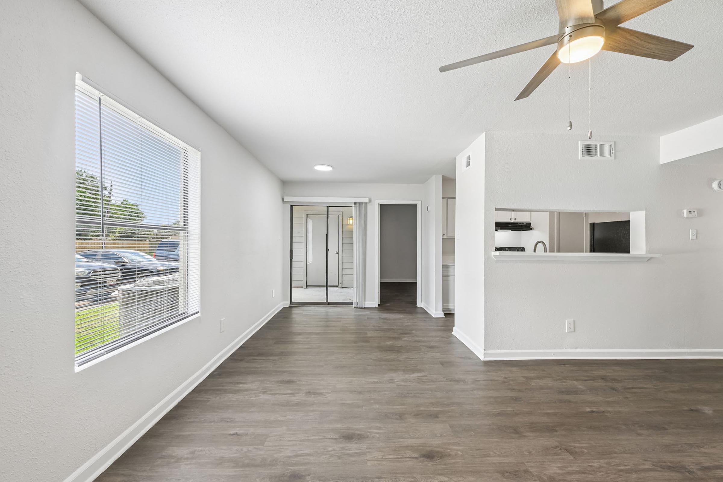 A bright and spacious living area with light-colored walls and wooden flooring. A ceiling fan circulates air, and large windows allow natural light in. The open layout includes a view of a nearby kitchen and a hallway leading to additional rooms. The overall ambiance is modern and inviting.