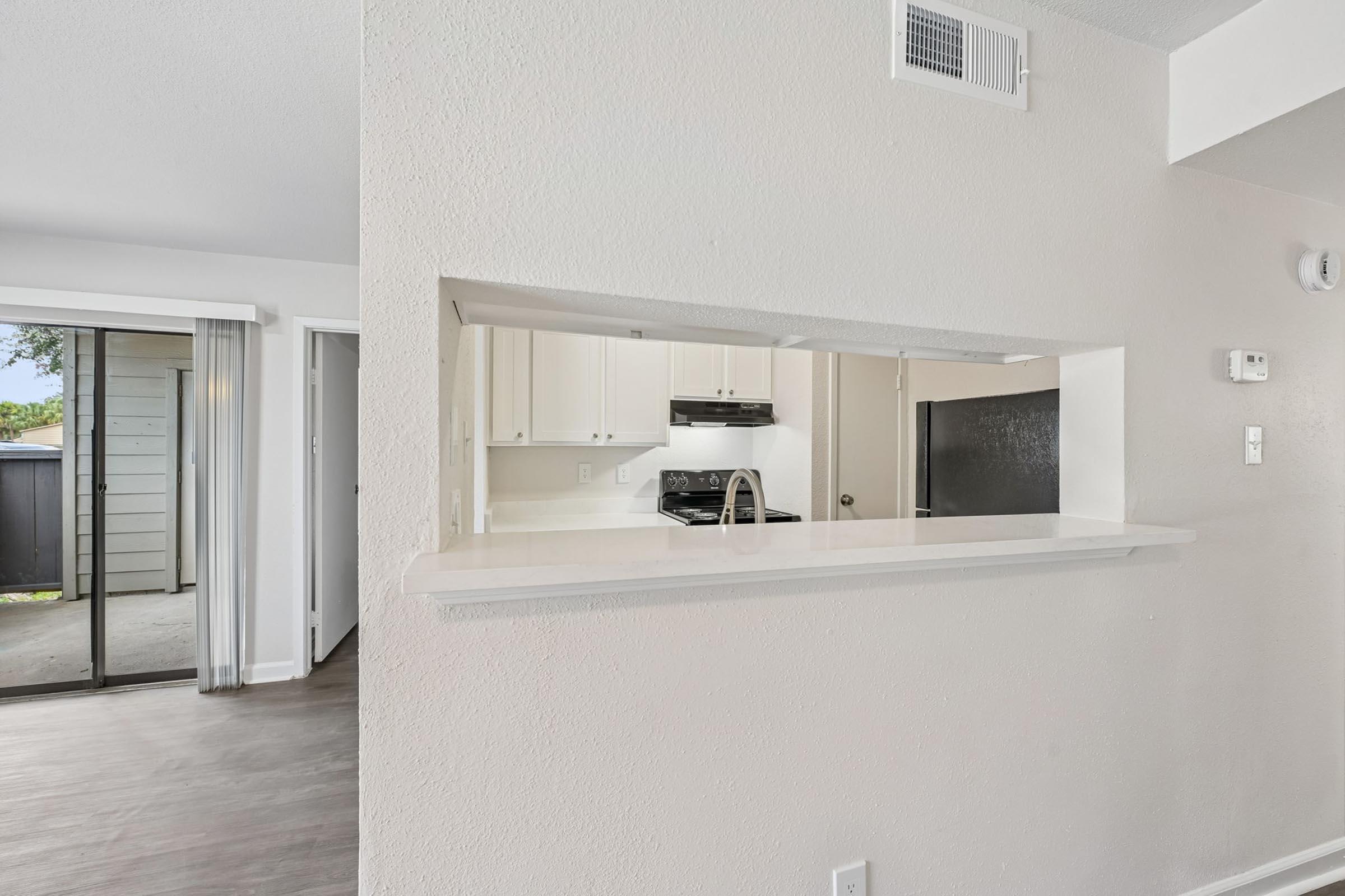 View from a living area into a kitchen through a pass-through countertop. The kitchen features white cabinetry and a black stove. Natural light comes from sliding glass doors leading to an outdoor patio area, which is partially visible. The space has neutral colors and modern design elements.