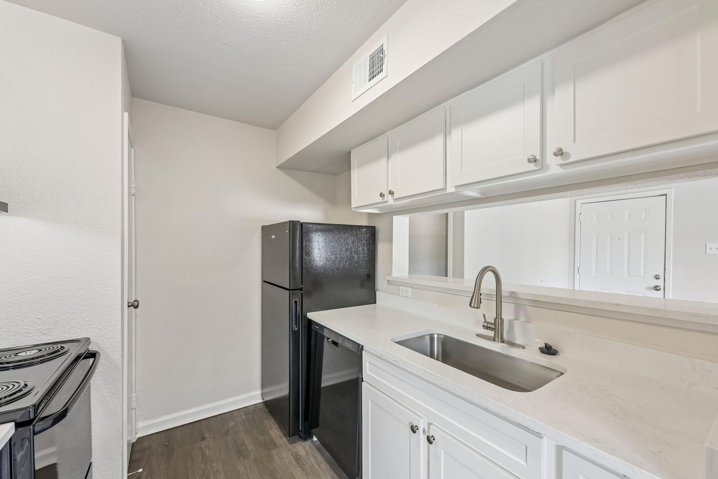 A modern kitchen featuring white cabinets, a black refrigerator, and a stainless steel sink. The counter is made of light-colored stone, and there's a stove visible. The walls are painted in a light color, creating a bright and clean atmosphere. A door leading to another room is visible in the background.