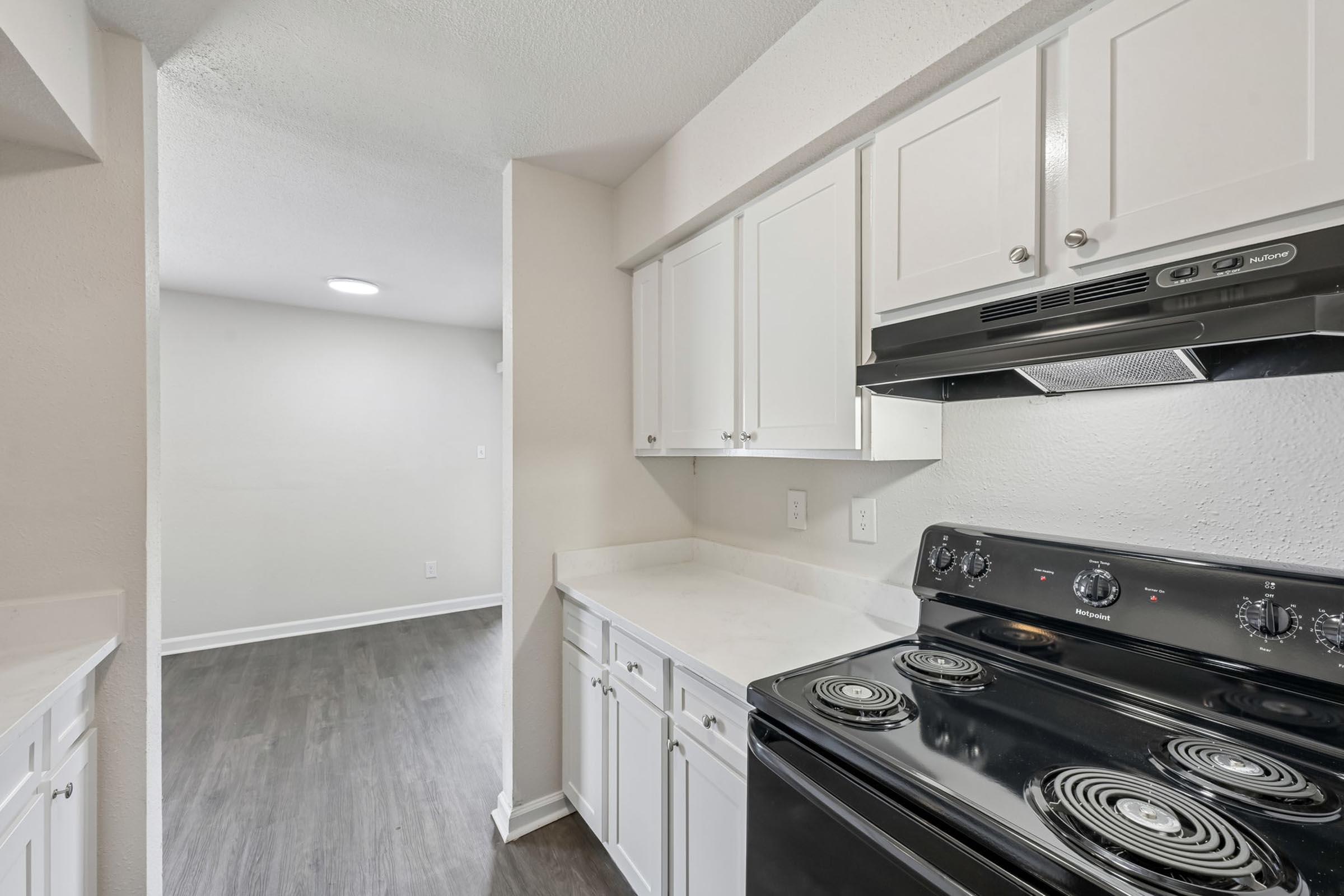 A modern kitchen featuring white cabinetry, a black stovetop and oven, and a clean countertop. The space transitions to a light-colored room in the background, with neutral walls and natural lighting. The overall aesthetic is bright and contemporary.