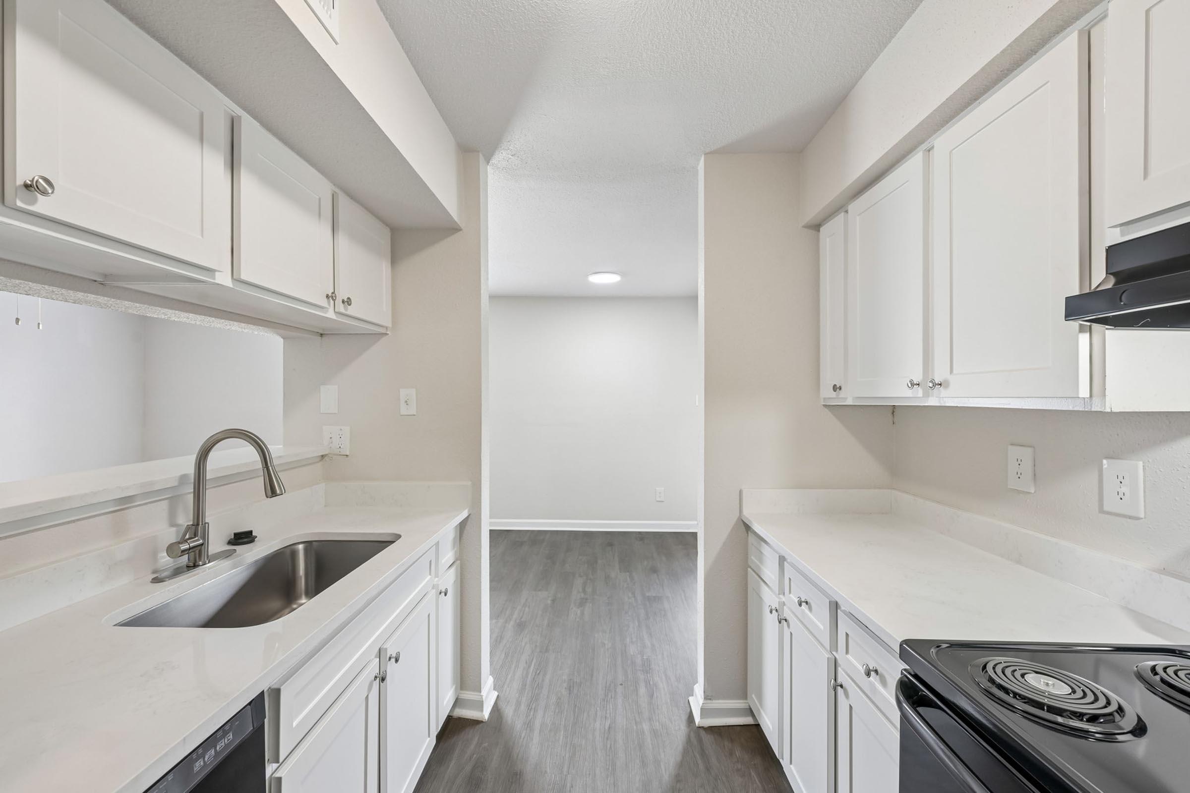 A modern kitchen featuring white cabinets, a stainless steel sink, and a black stove. The countertops are light-colored, and the flooring is a laminate in a gray shade. The layout opens to a bright living space with natural light and neutral walls.