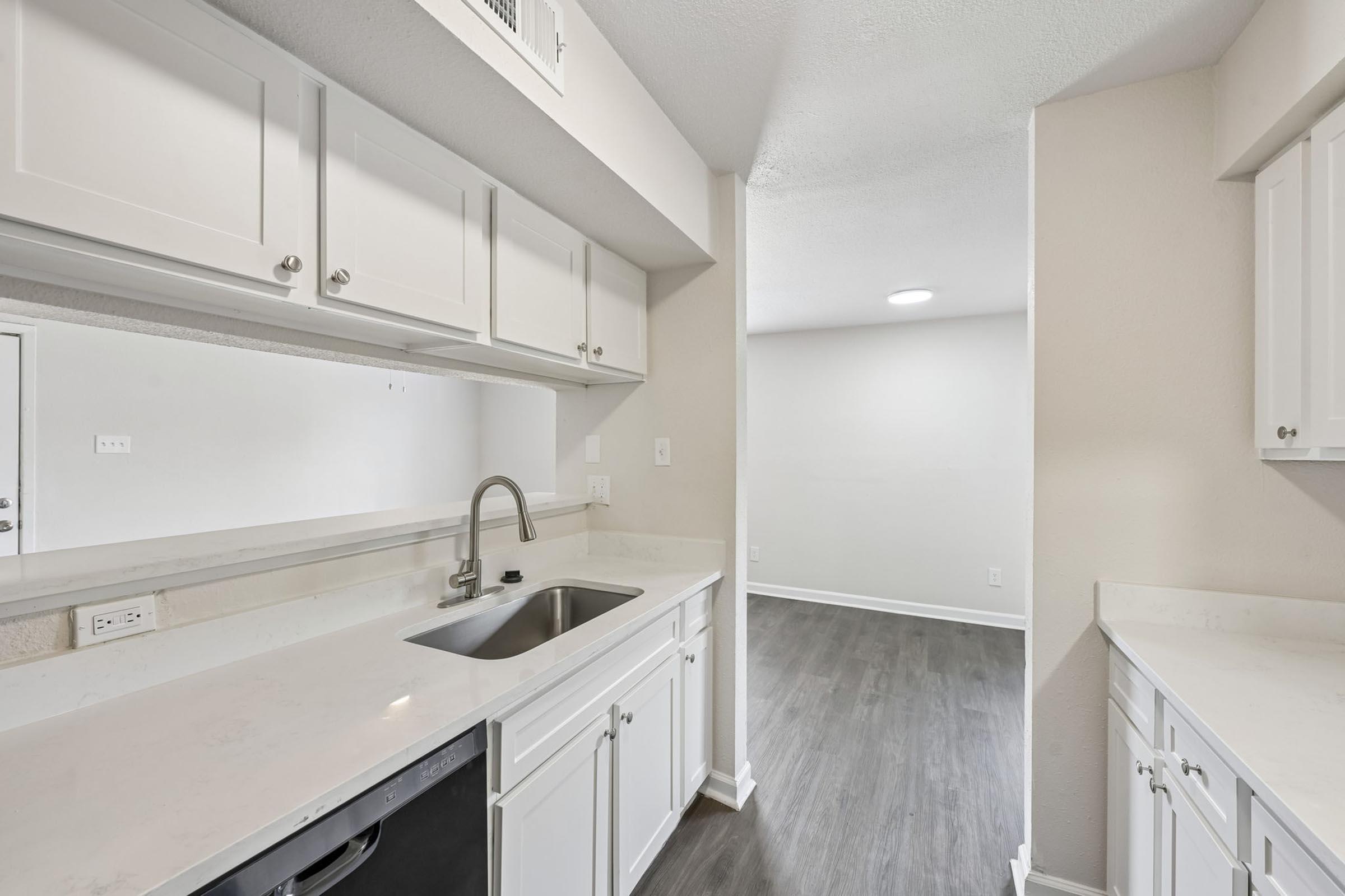 Modern kitchen with white cabinetry and a stainless steel sink, featuring a clean, minimalist design. There is a dishwasher under the counter and smooth, light-colored countertops. The background shows an adjacent room with light-colored walls and a neutral floor, enhancing the open and airy feel of the space.