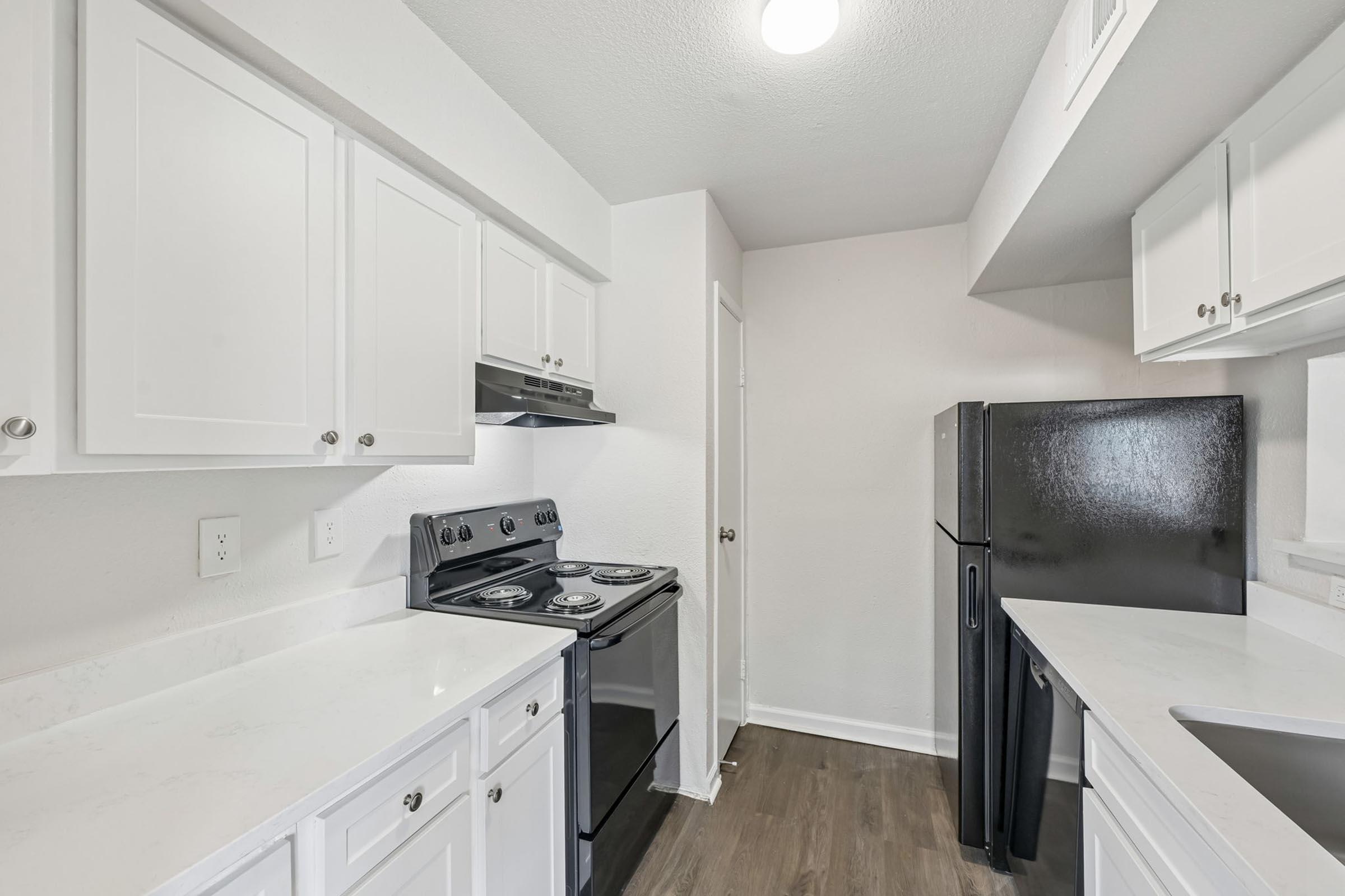 A modern kitchen featuring white cabinetry, a black stove, and a black refrigerator. The countertops are light-colored, and the space is well-lit with a ceiling light. The kitchen has an open layout with a door leading to another room.