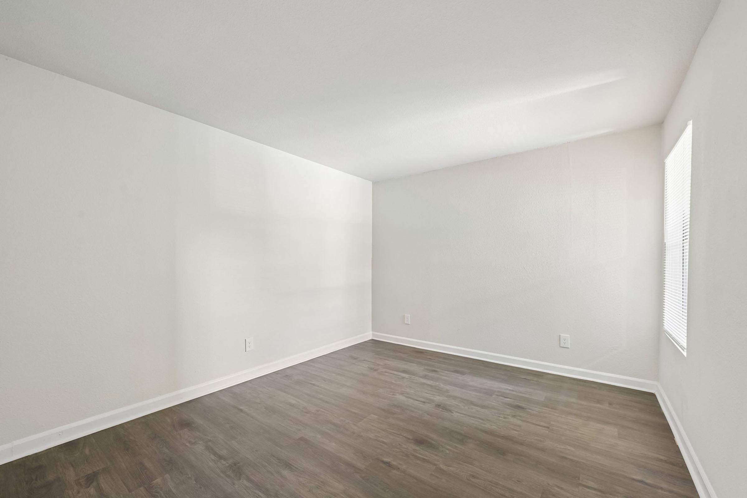 Empty room with light gray walls and dark wood flooring. One small window on the right wall allows natural light into the space. The room has a clean and minimalistic appearance, ideal for various uses.