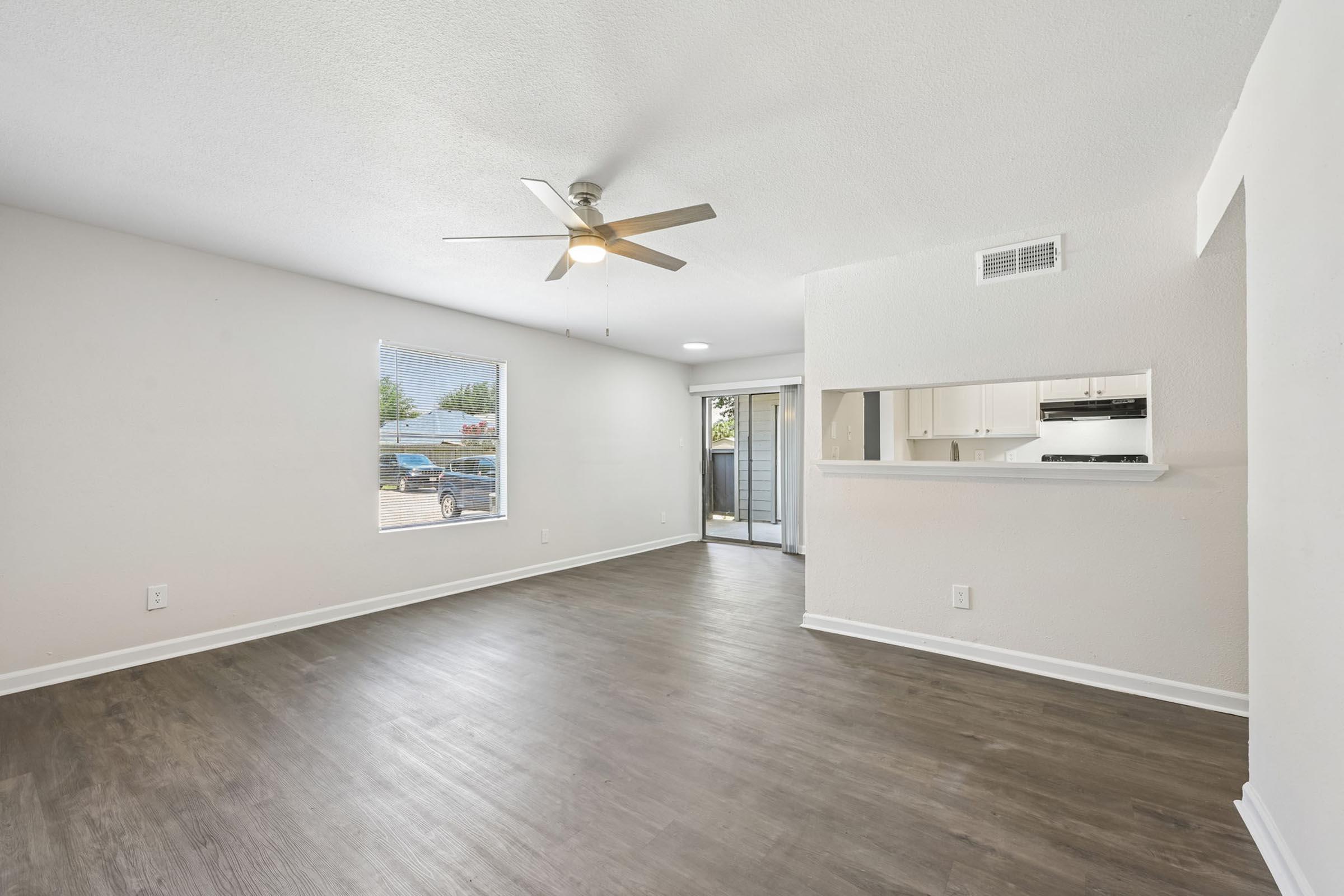 Spacious, modern living room with light-colored walls and a ceiling fan. The room features large windows allowing natural light, light hardwood flooring, and an open layout leading to a kitchen area. A sliding glass door opens to an outdoor space, enhancing the airy feel of the home.
