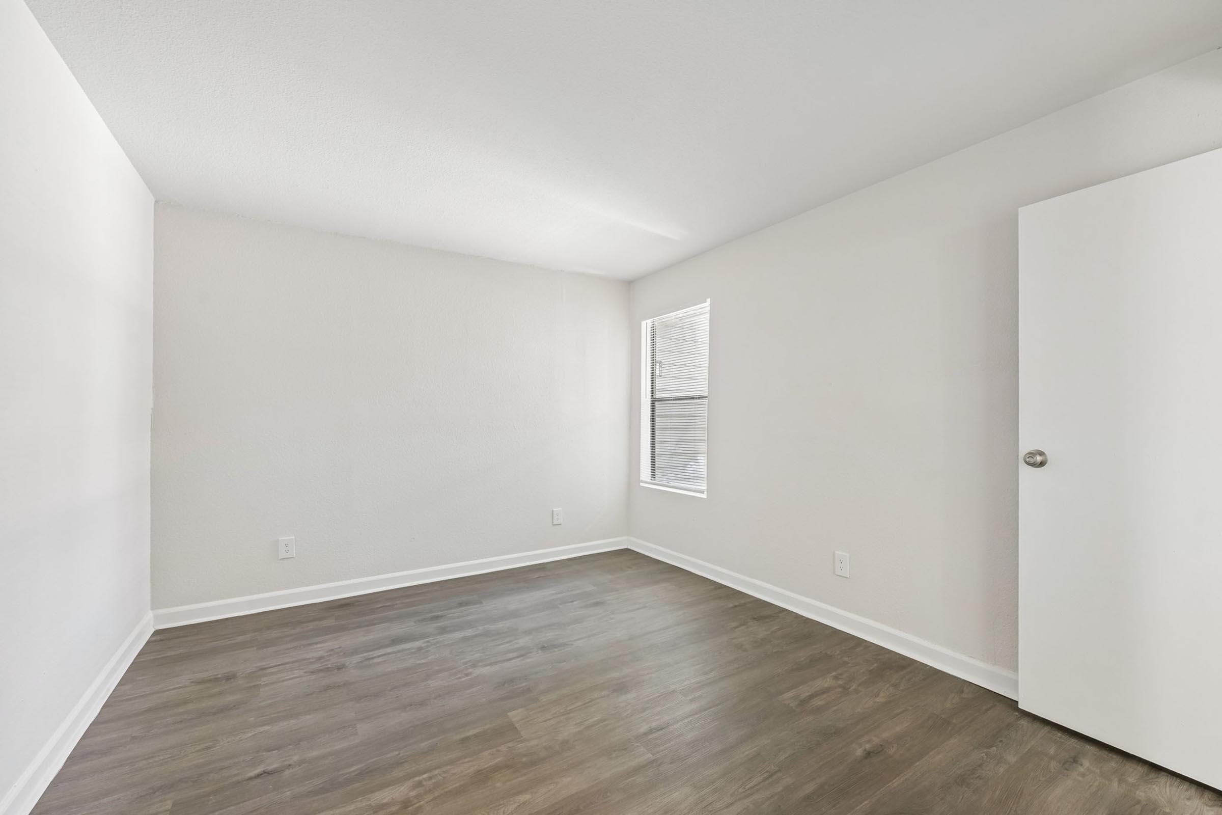 A spacious, empty room featuring light-colored walls and a wide window allowing natural light. The floor is a light wood laminate, and there is a closed white door on the right. The overall atmosphere is bright and minimalistic, making it suitable for various uses or decoration.