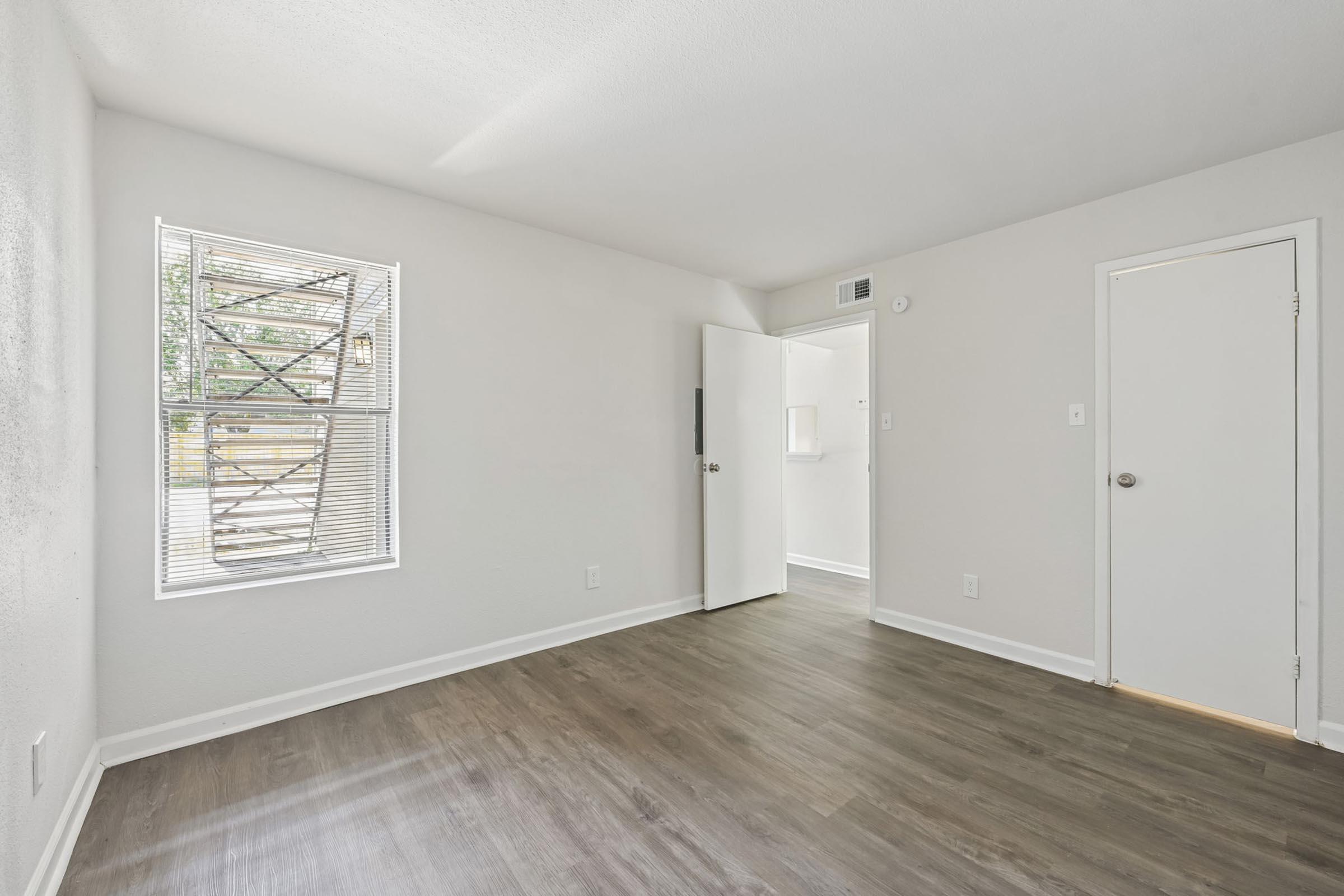 Empty room with light gray walls and a wood-like floor. A window with blinds allows natural light in, while a door leads to another room. The space appears clean and modern, suitable for various uses.