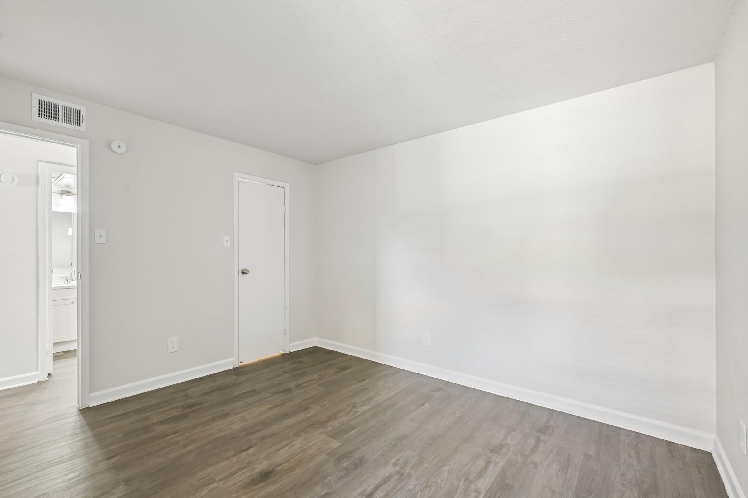 A light-filled, empty room with a smooth, painted white wall and a wooden floor. There is a door on the left leading to a bathroom and another door to the right. The minimalist space has no furniture, creating a clean and open atmosphere, ideal for a living area or bedroom.