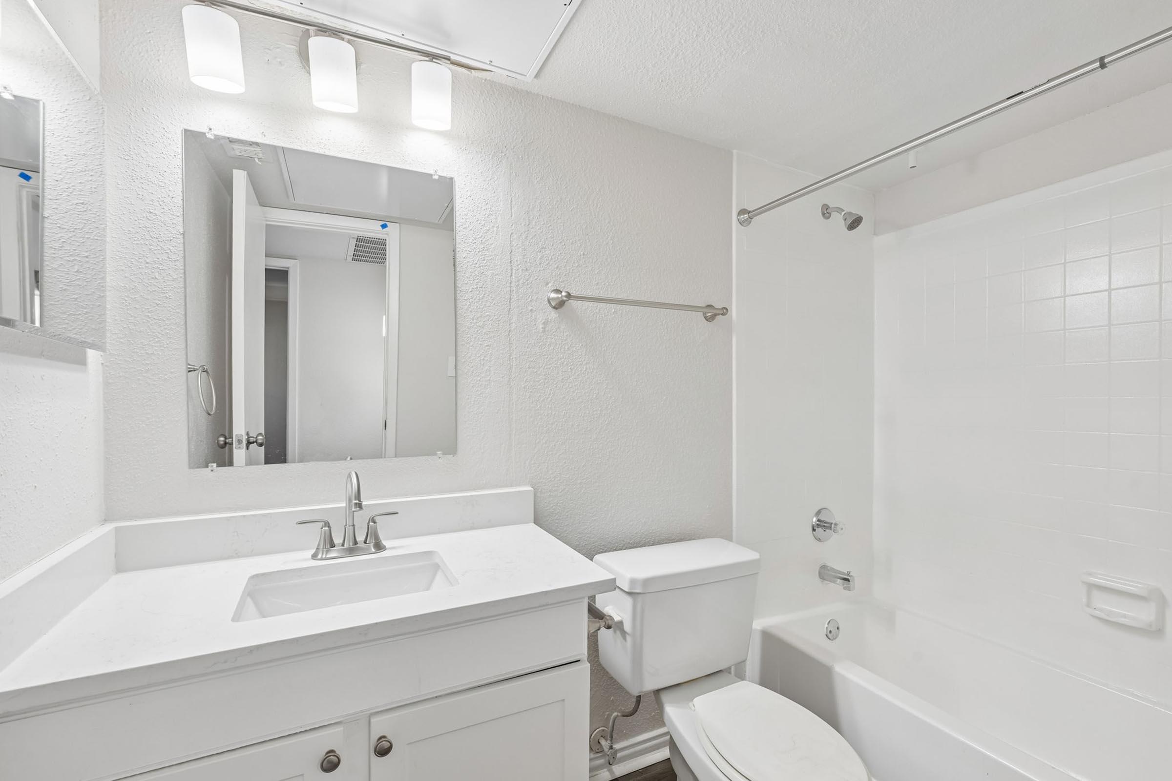 A modern bathroom featuring a white vanity with a sink, a large mirror above it, a toilet, and a bathtub with a shower. The walls are light-colored, and there is overhead lighting with three fixtures. The bathroom appears bright and clean.