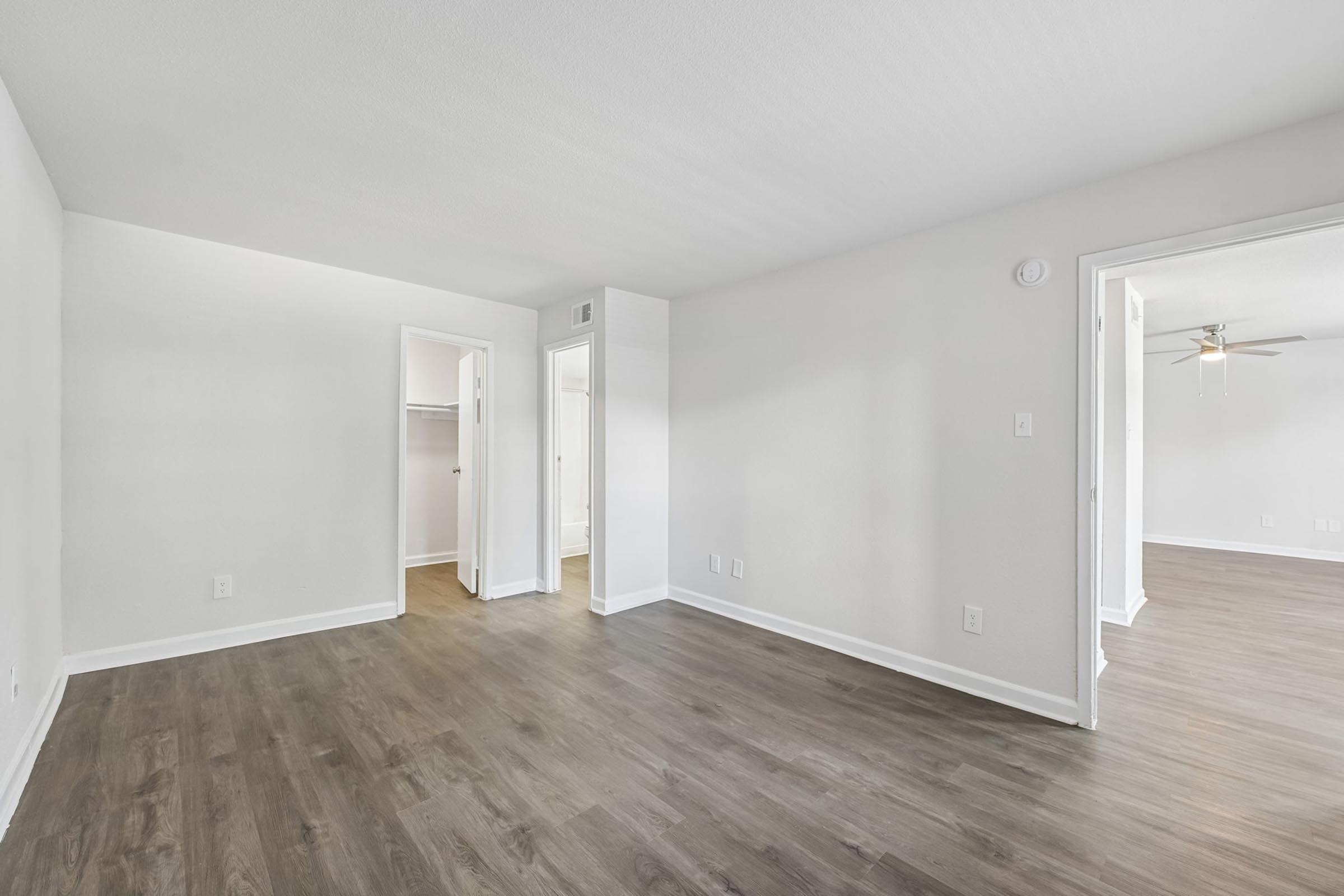 A spacious, empty room with light-colored walls and a light wood floor. There are two doorways leading to other rooms, one of which has a small closet. The ceiling features a ceiling fan, and natural light comes in from the open doorway on the right. The overall atmosphere is bright and inviting.