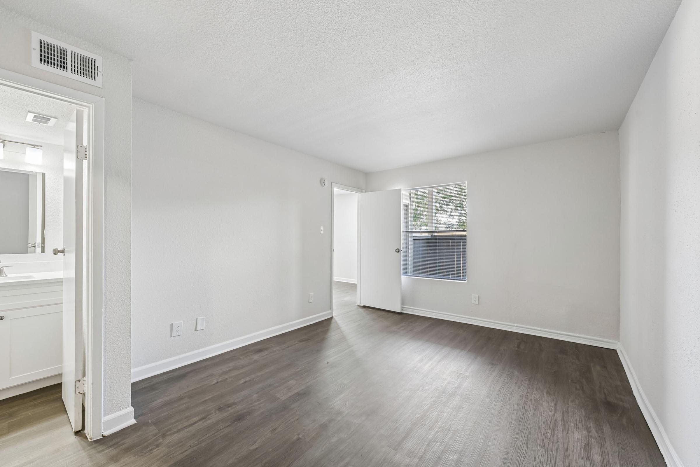 A spacious, empty room featuring light-colored walls and a smooth floor. There is a doorway leading to an adjacent area, and a window allows natural light to enter. The overall feel is bright and clean, suitable for various uses such as a living space or office.