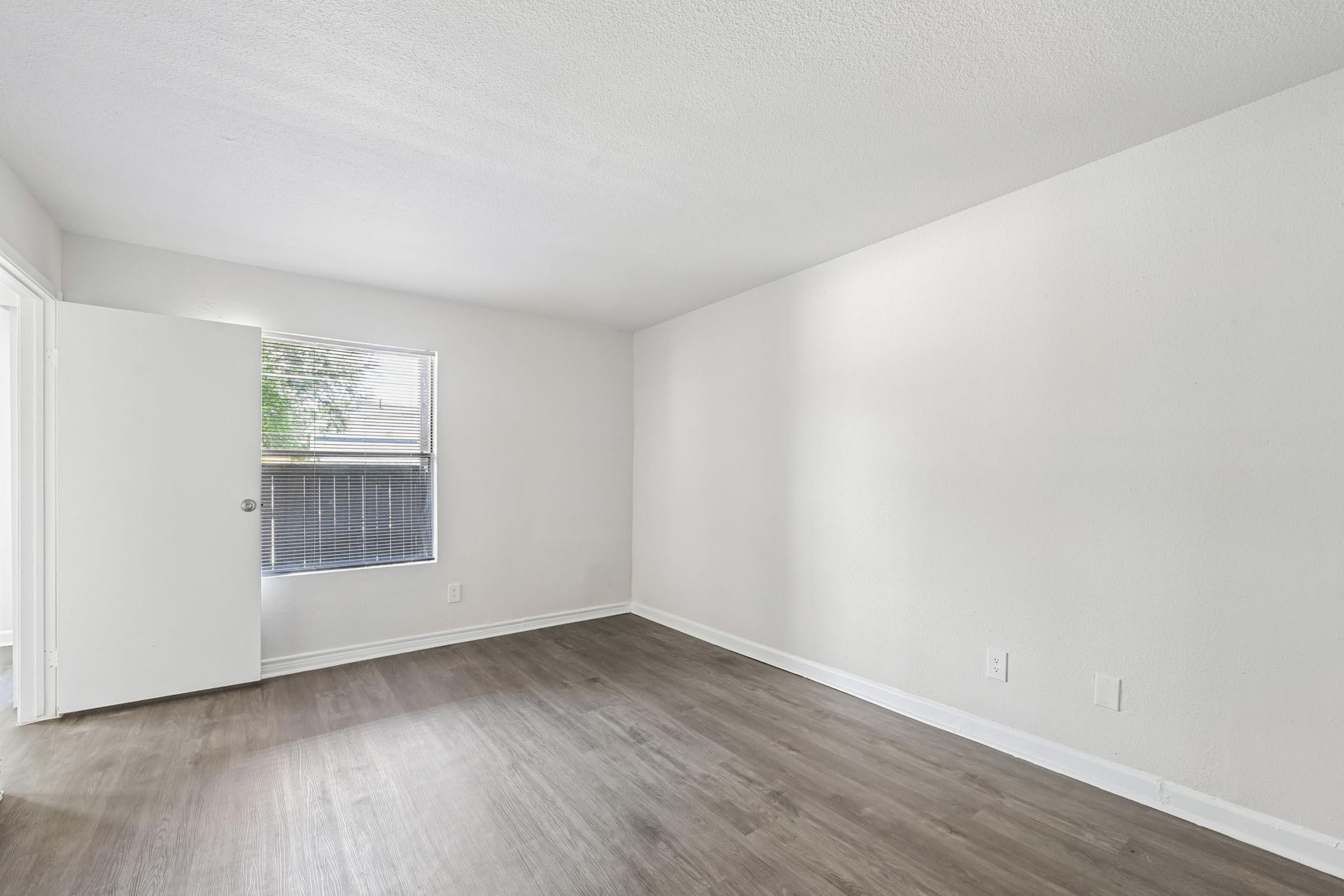 Empty room with light-colored walls and a wooden floor. A window with blinds allows natural light to enter from the left side, and a partially open door is visible to the left. The room is unfurnished, giving it a spacious and clean appearance.