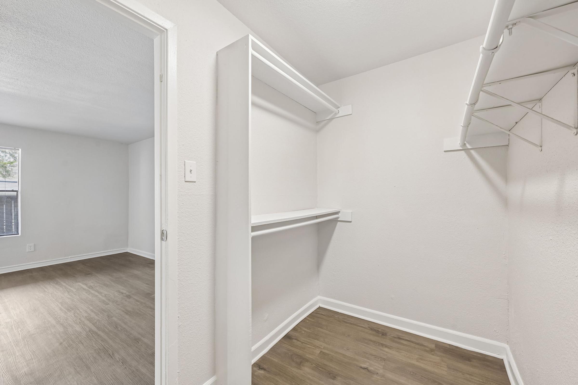 A spacious room with light-colored walls and a wooden floor. On the left, there is an empty closet featuring built-in shelves and a hanging rod. The doorway opens to another area, possibly a living space, visible in the background.