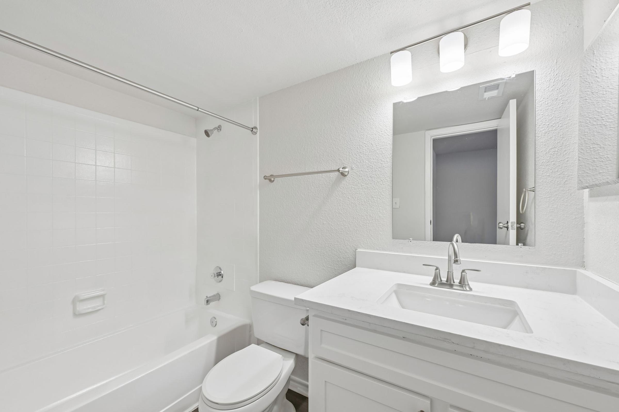 A clean, modern bathroom featuring a white bathtub with a shower, a sleek sink with a chrome faucet, white cabinetry, and a large mirror. Bright overhead lighting illuminates the space, and a door is partially visible in the background leading to another room.