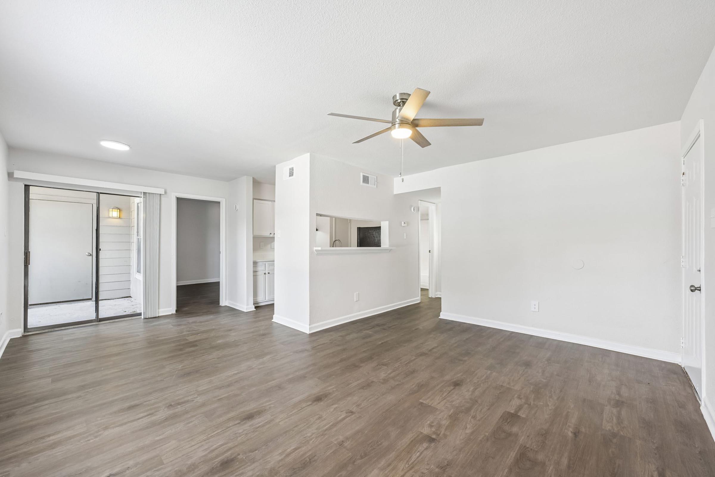 Bright and spacious living area featuring wood-style flooring, a ceiling fan, and an open layout. It includes a view of a sliding glass door leading to an outdoor space, along with access to a kitchen area. Walls are painted in a light color, enhancing the airy feel of the room.