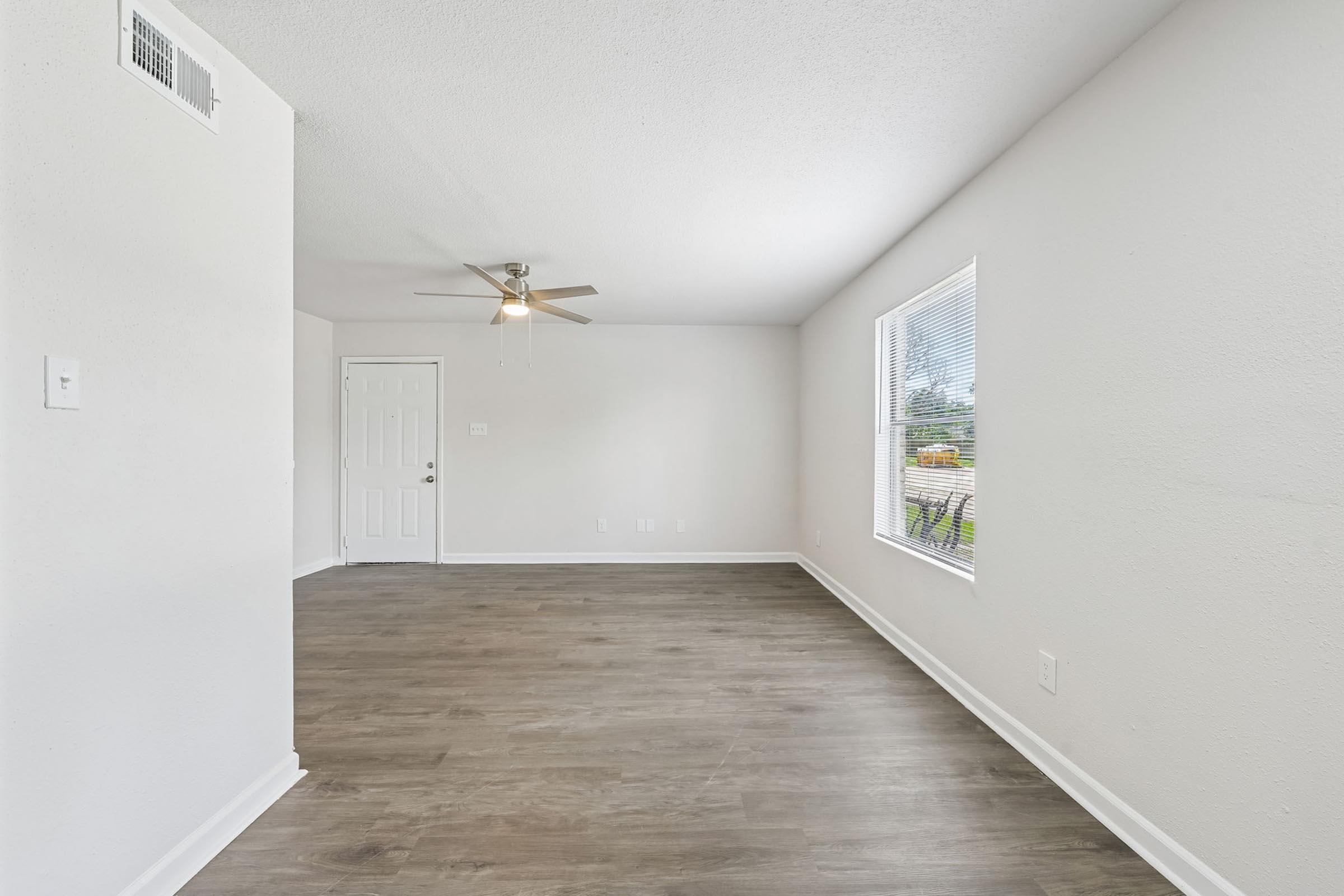 A bright, vacant interior room with light-colored walls and a ceiling fan. The floor is covered with light wood laminate. There is a door visible on the left leading to another room, and a window on the right letting in natural light. The space is clean and sparsely furnished.
