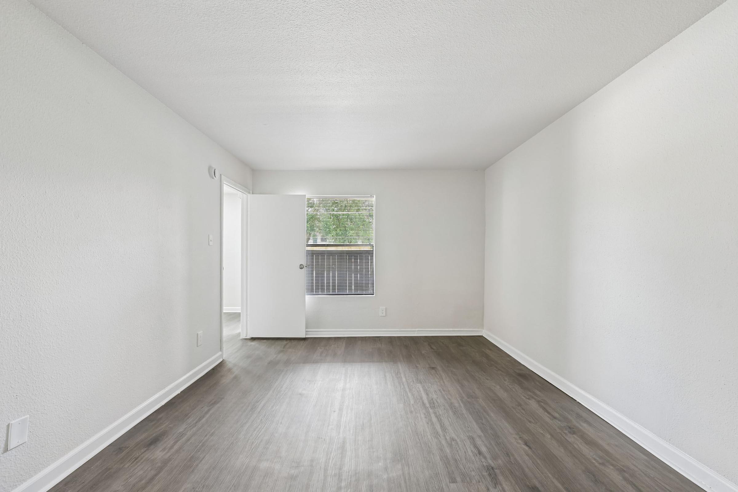 Empty room with light-colored walls and a smooth, dark wood-style floor. A window allows natural light to enter, and a partially open door leads to another space. The room has a clean, minimalist aesthetic, making it suitable for various uses or decoration.