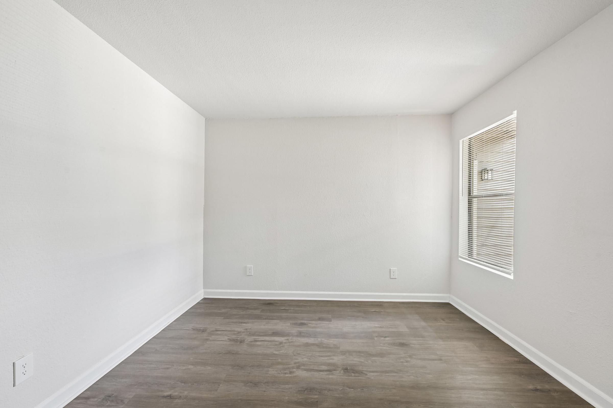 Empty room with light gray walls and hardwood flooring. One window with blinds allows natural light to enter. The space is minimalistic and unadorned, creating a calm and open atmosphere.