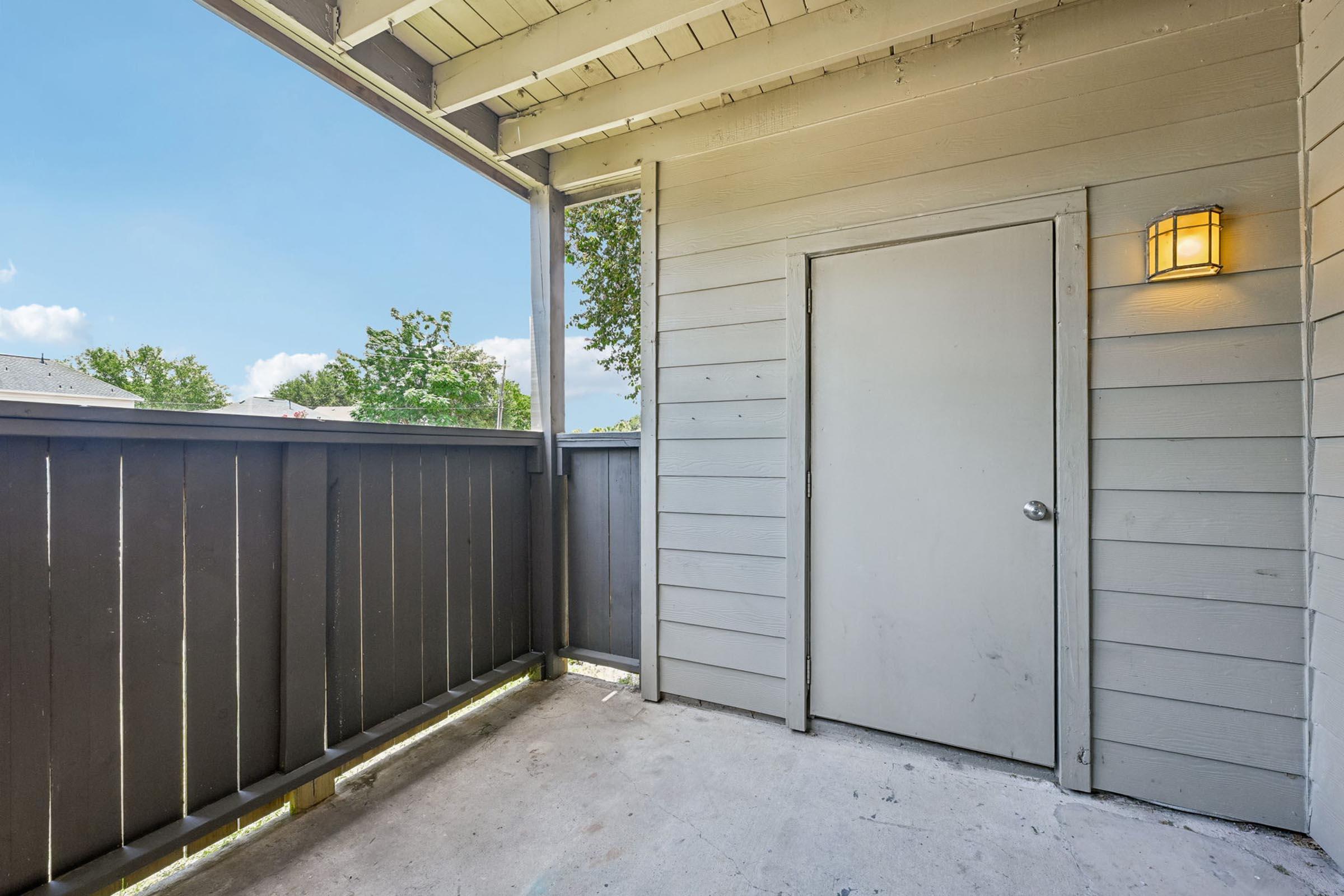 A small outdoor balcony or porch featuring a gray wooden railing, a plain white door, and a wall-mounted light fixture. The area is partially shaded with a blue sky and trees visible in the background. The floor is bare concrete, and the space is uncluttered and minimalistic.