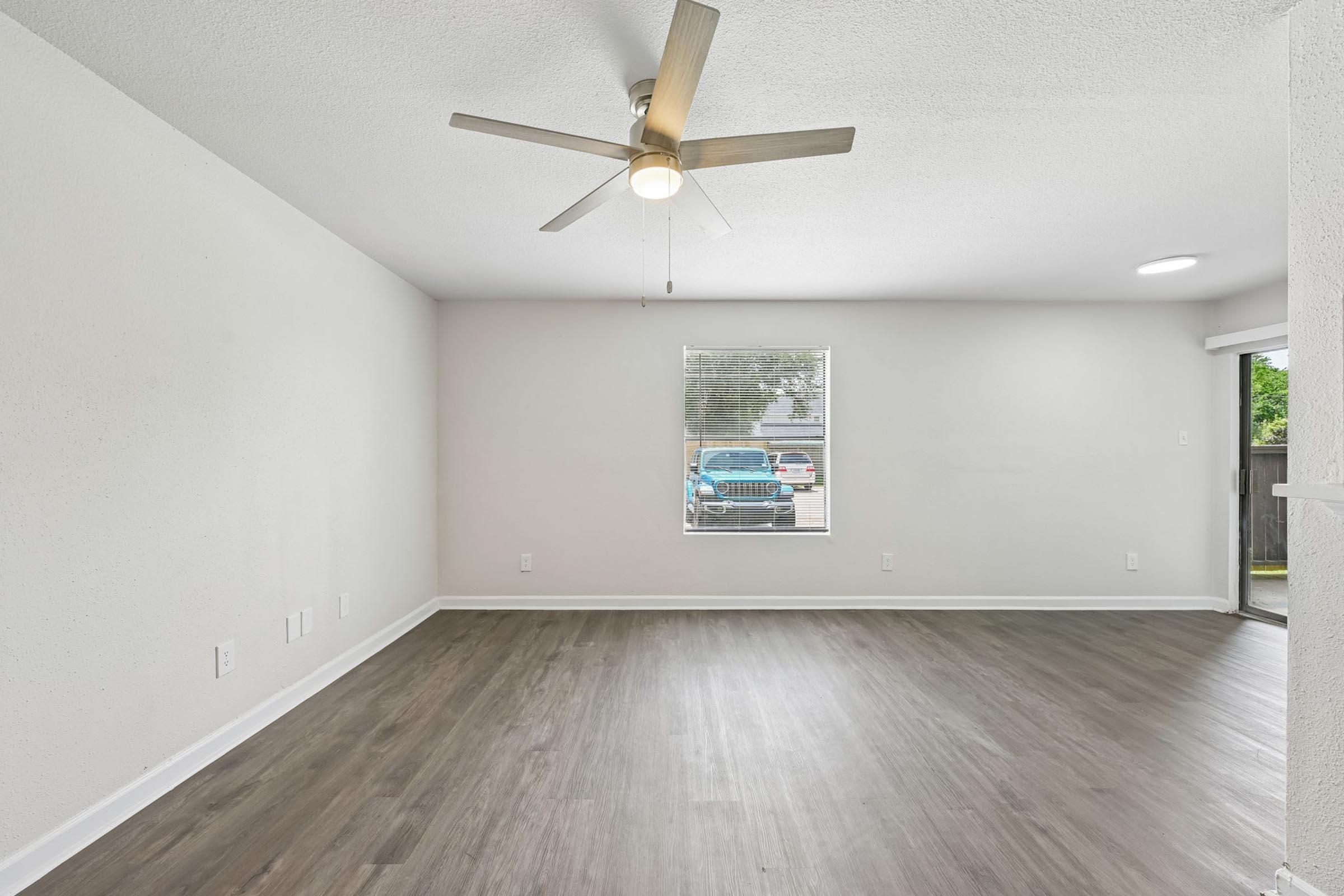 A well-lit, empty living room featuring light gray walls and a ceiling fan. The floor is made of light wood laminate. A window in the background shows a glimpse of a blue vehicle outside. A doorway leads to another space in the room. The atmosphere is open and airy, creating a spacious feel.