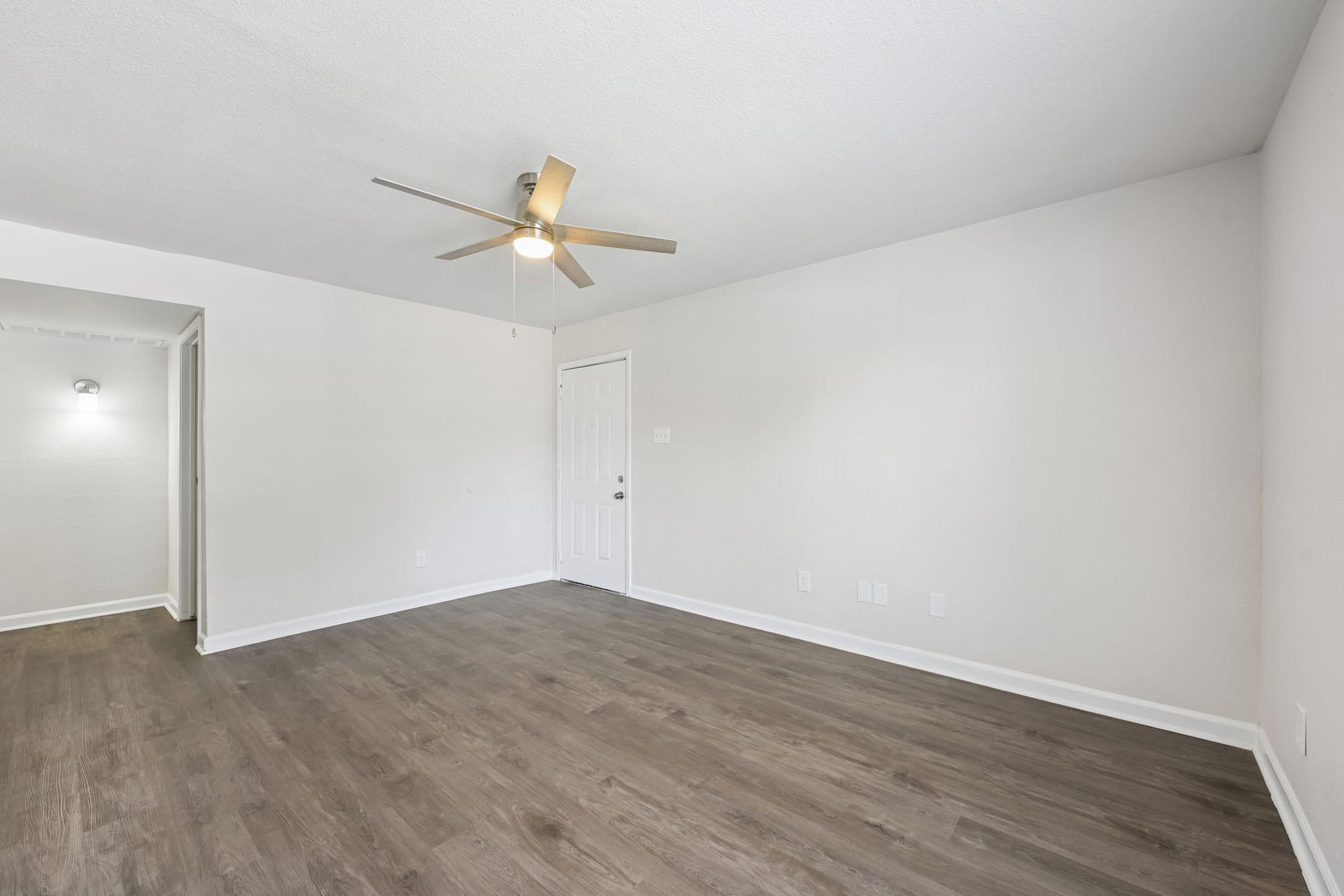 A spacious, empty room with light-colored walls and a ceiling fan. The flooring is a modern laminate, and there's a door leading outside. Minimalistic decor creates a bright, airy atmosphere, making it suitable for various uses.