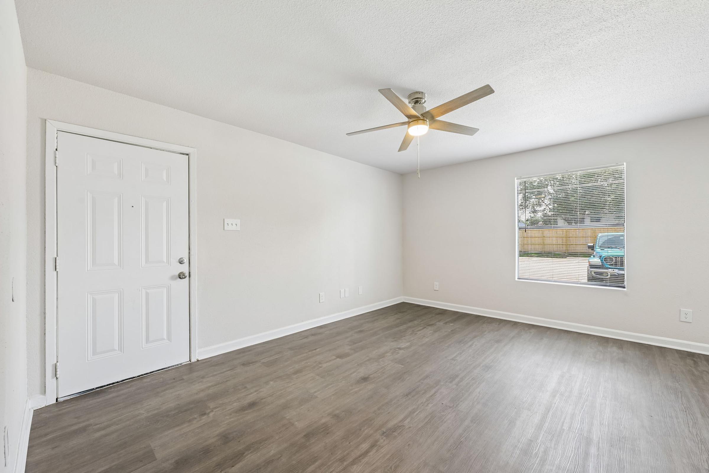 A spacious, empty room with light-colored walls and laminate flooring. A ceiling fan is mounted in the center. To the left, there is a white door leading outside, and a large window on the right allows natural light to enter, illuminating the space. A vehicle is visible outside through the window.