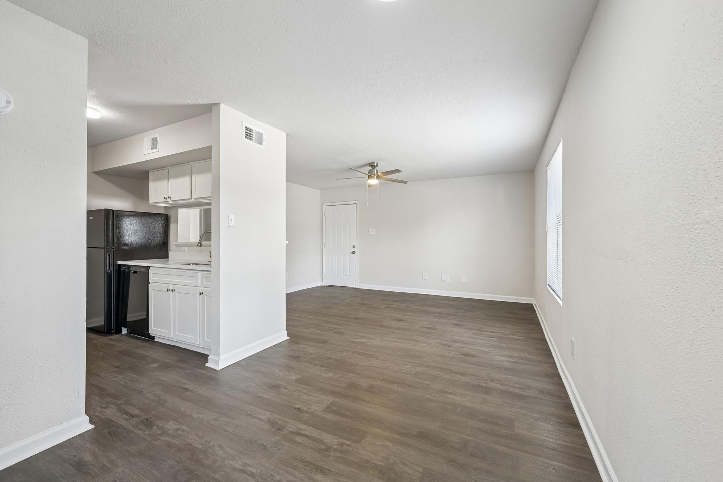 A spacious, empty living area featuring light-colored walls and wood-style flooring. An open kitchen with white cabinetry and a black refrigerator is visible on the left. The room includes a ceiling fan and a door leading to another area, with windows allowing natural light to enter.