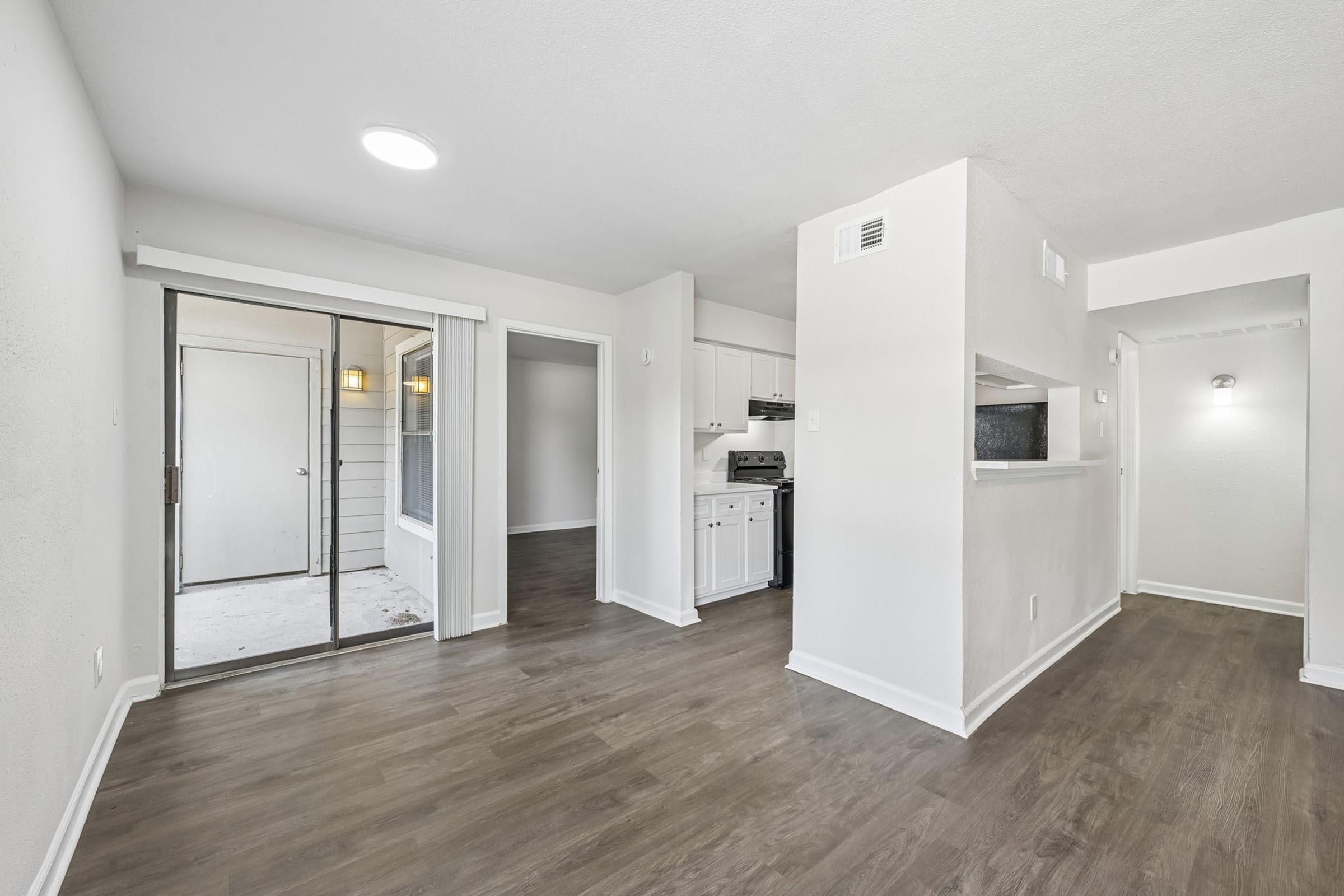 Interior view of a modern apartment featuring a spacious living area, light-colored walls, and laminate flooring. There is a sliding glass door leading to an outdoor space, and a partial wall dividing the living area from the kitchen. Natural light is present, creating a bright and inviting atmosphere.