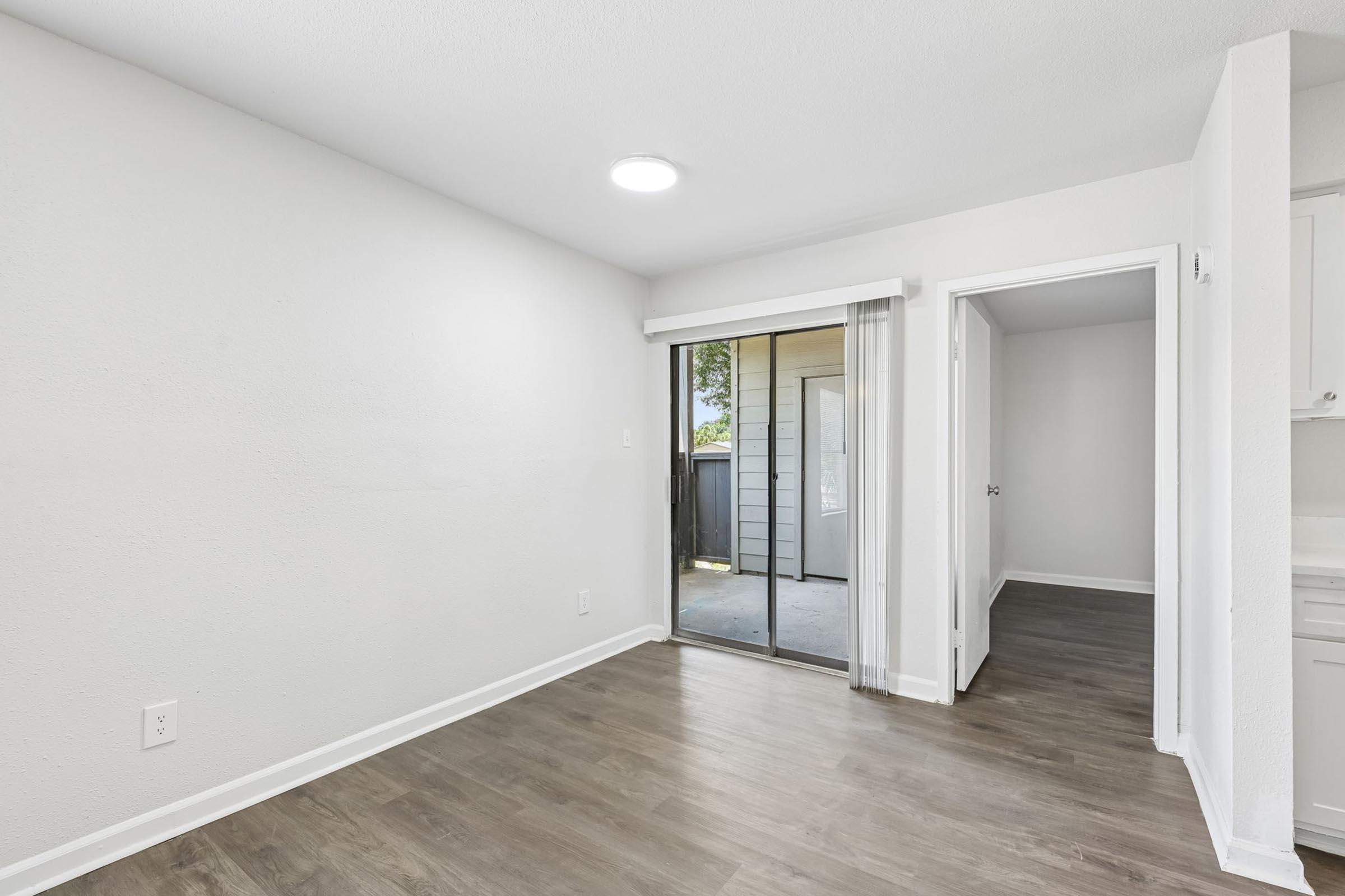 A bright, empty interior space featuring light-colored walls and laminate flooring. On the left, a sliding glass door leads to an outdoor area, while there is an open doorway on the right leading to another room. The space is well-lit with natural light and has a clean, modern feel.