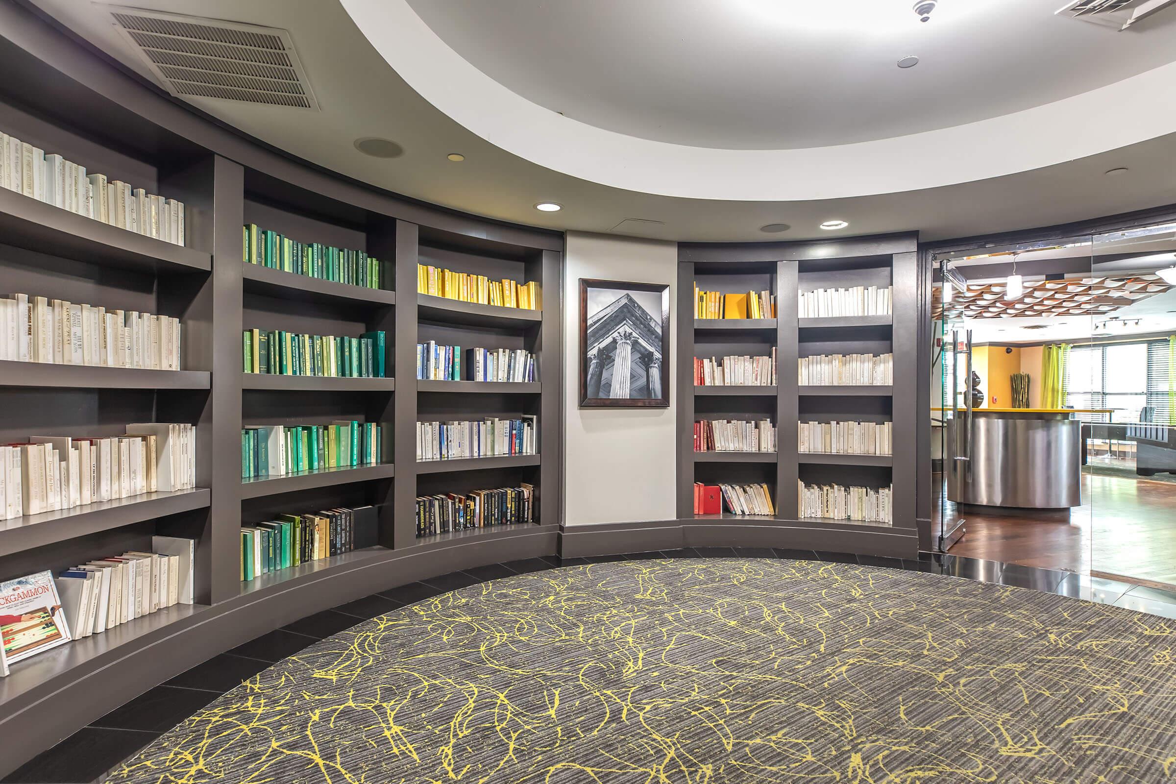 Curved bookcase filled with neatly arranged books in various colors, including white, green, and yellow. The backdrop features a modern interior with a circular carpet and an artistic light fixture. A framed black-and-white photograph of an architectural element can be seen on the wall.
