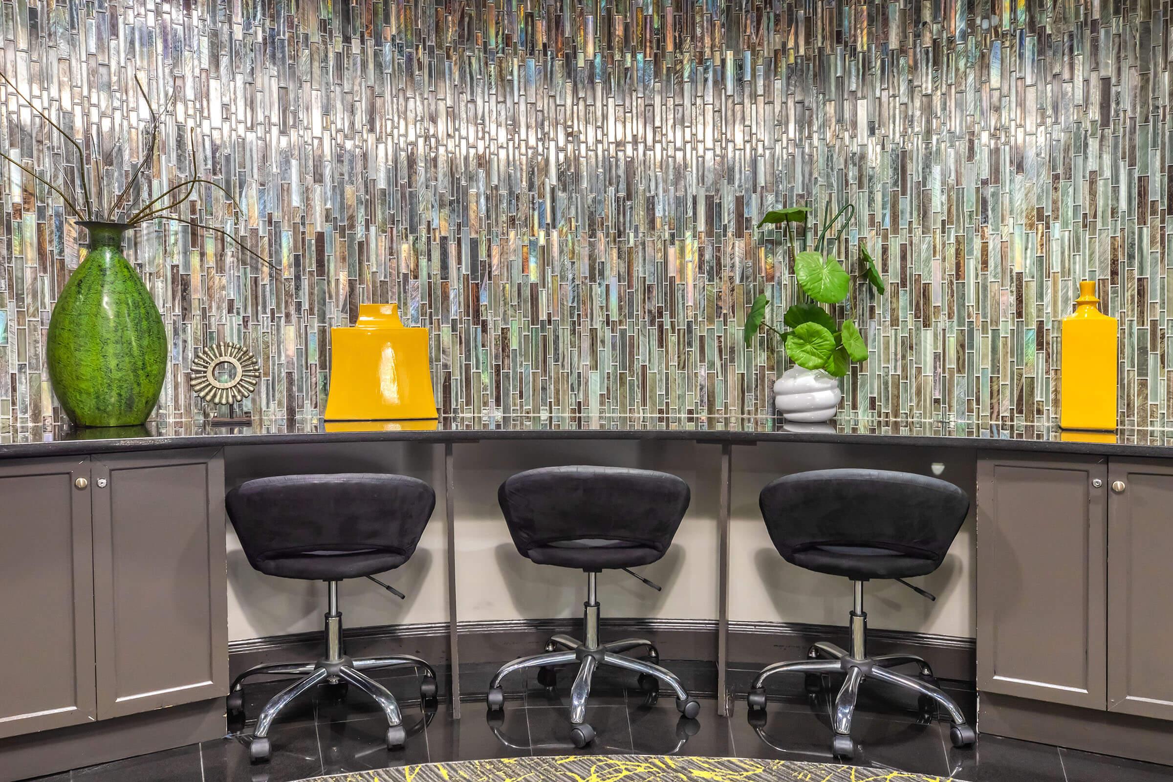 A stylish interior space featuring a bar-like counter with three black swivel chairs. Behind the counter is a colorful, textured mosaic wall made of glass tiles. Decor elements include a large green vase, a bright yellow pitcher, and a white planter with greenery.